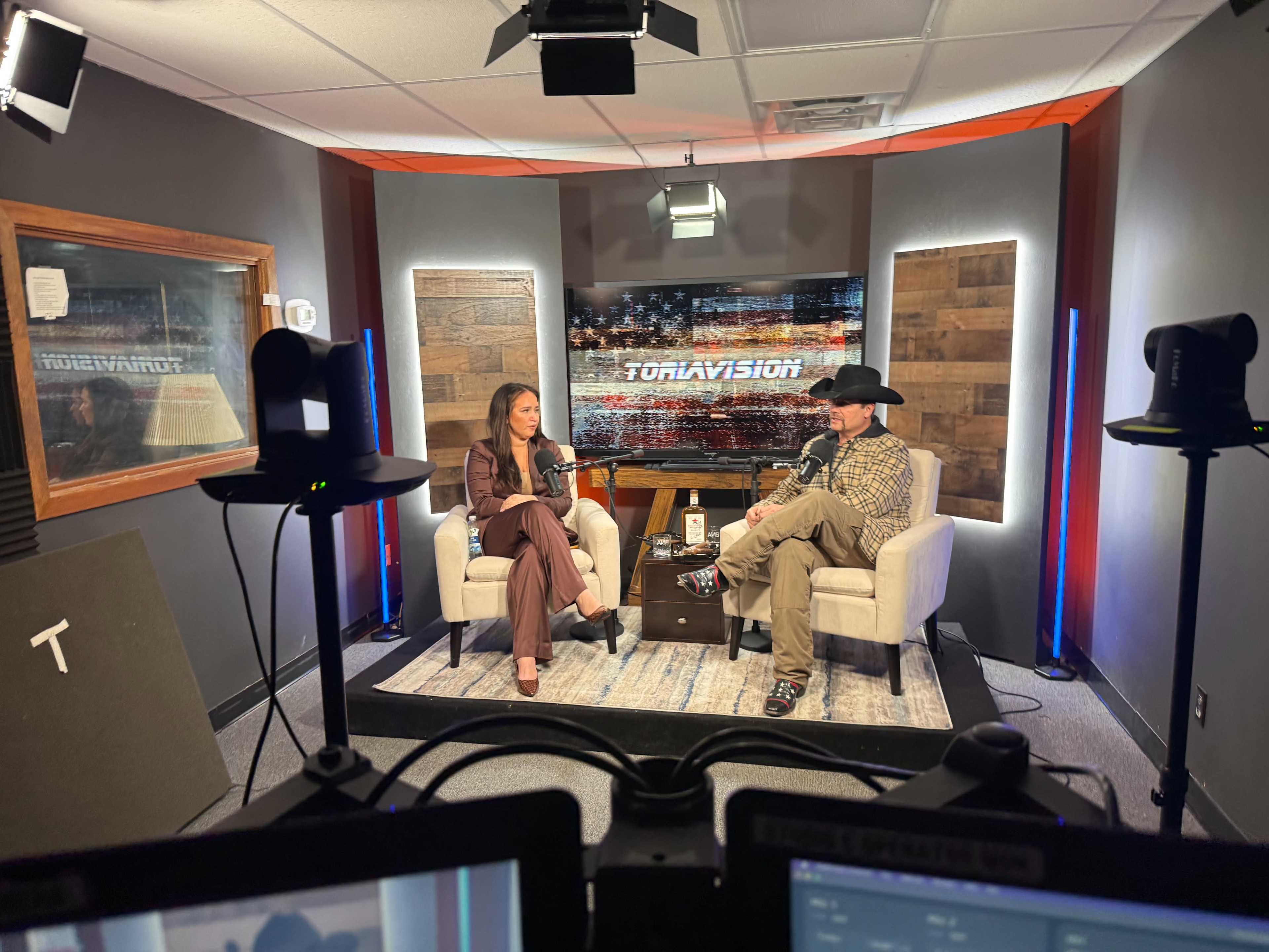 A woman and a man in a cowboy hat sit in armchairs on a television set, discussing in front of a backdrop displaying the word "TORAVISION."