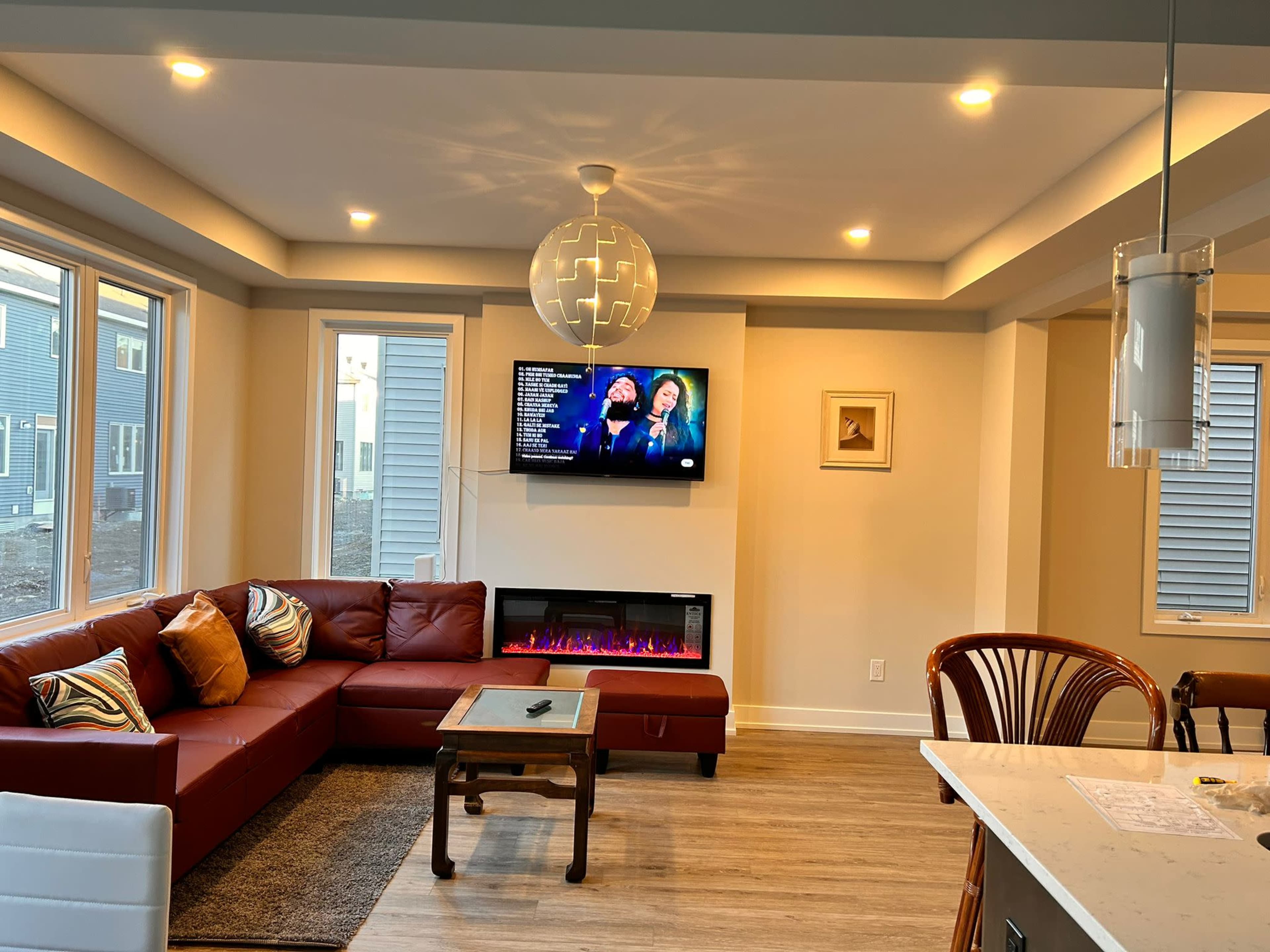 A modern living room featuring a red sectional couch, a wall-mounted television, a decorative light fixture, and an electric fireplace beneath a large window.