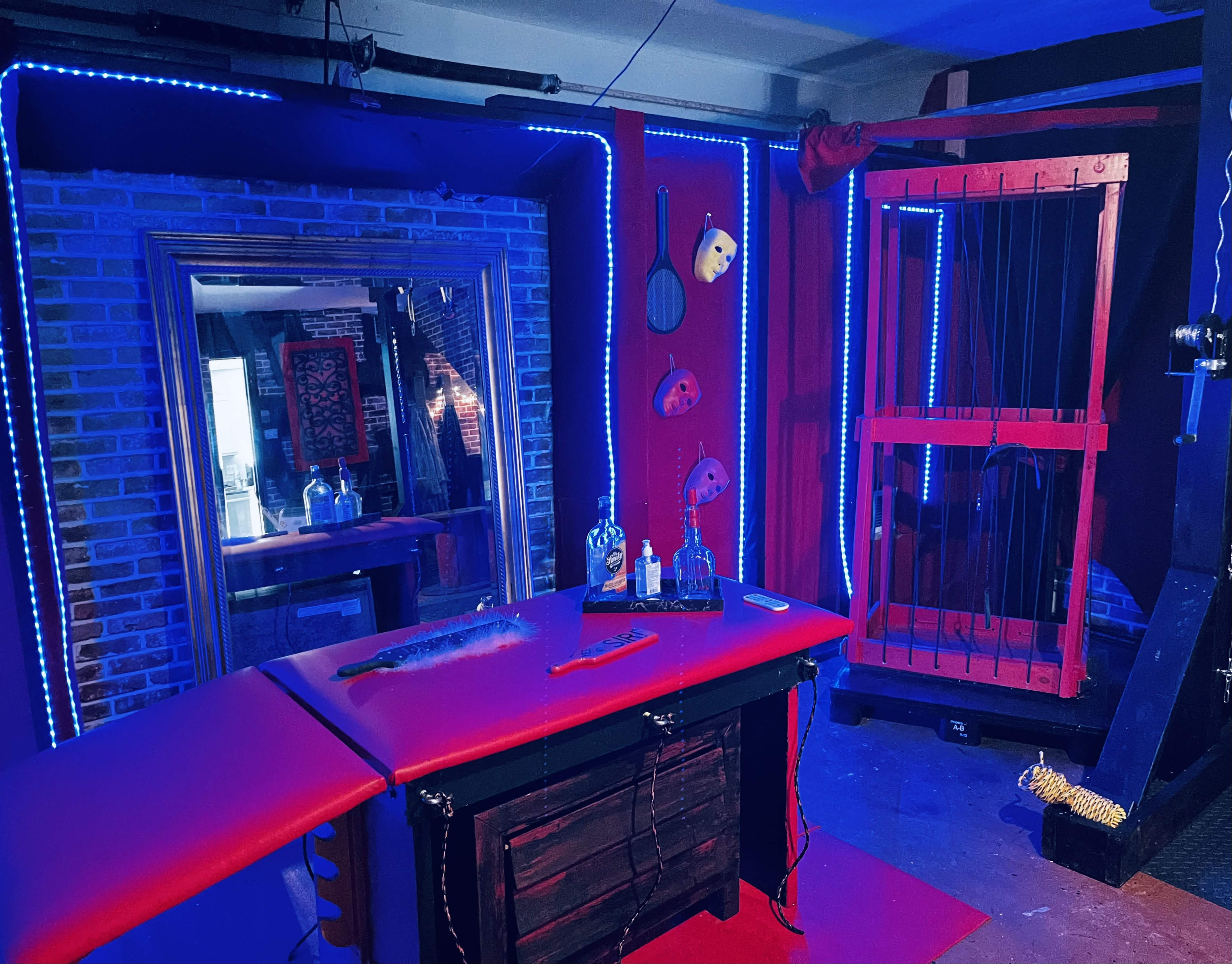 A dimly lit room featuring a brick wall, a large mirror, a red table with various tools, and a cage on the side, illuminated by blue LED lights.