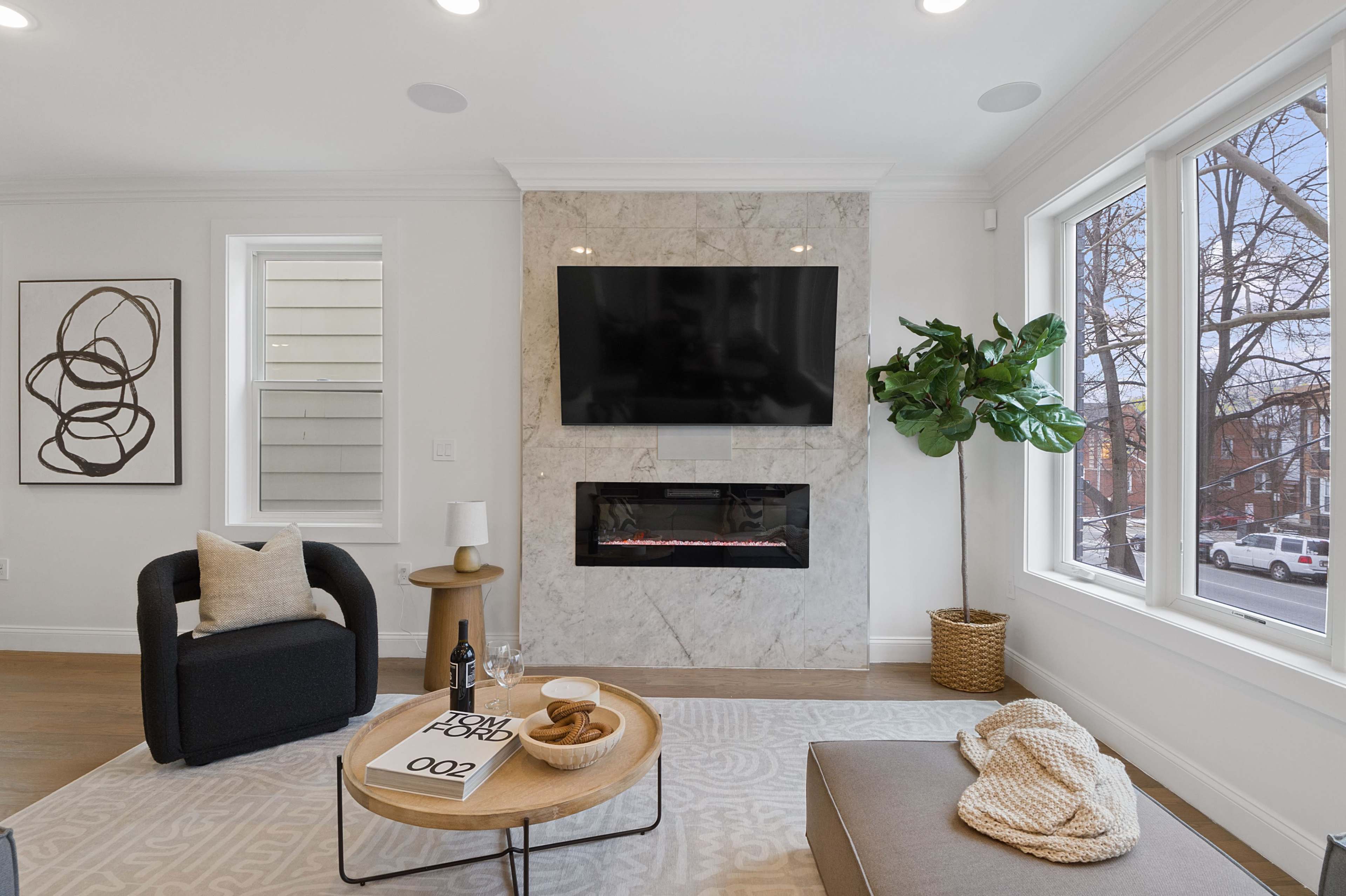 A modern living room features a mounted television above a sleek fireplace, with large windows and contemporary furnishings.