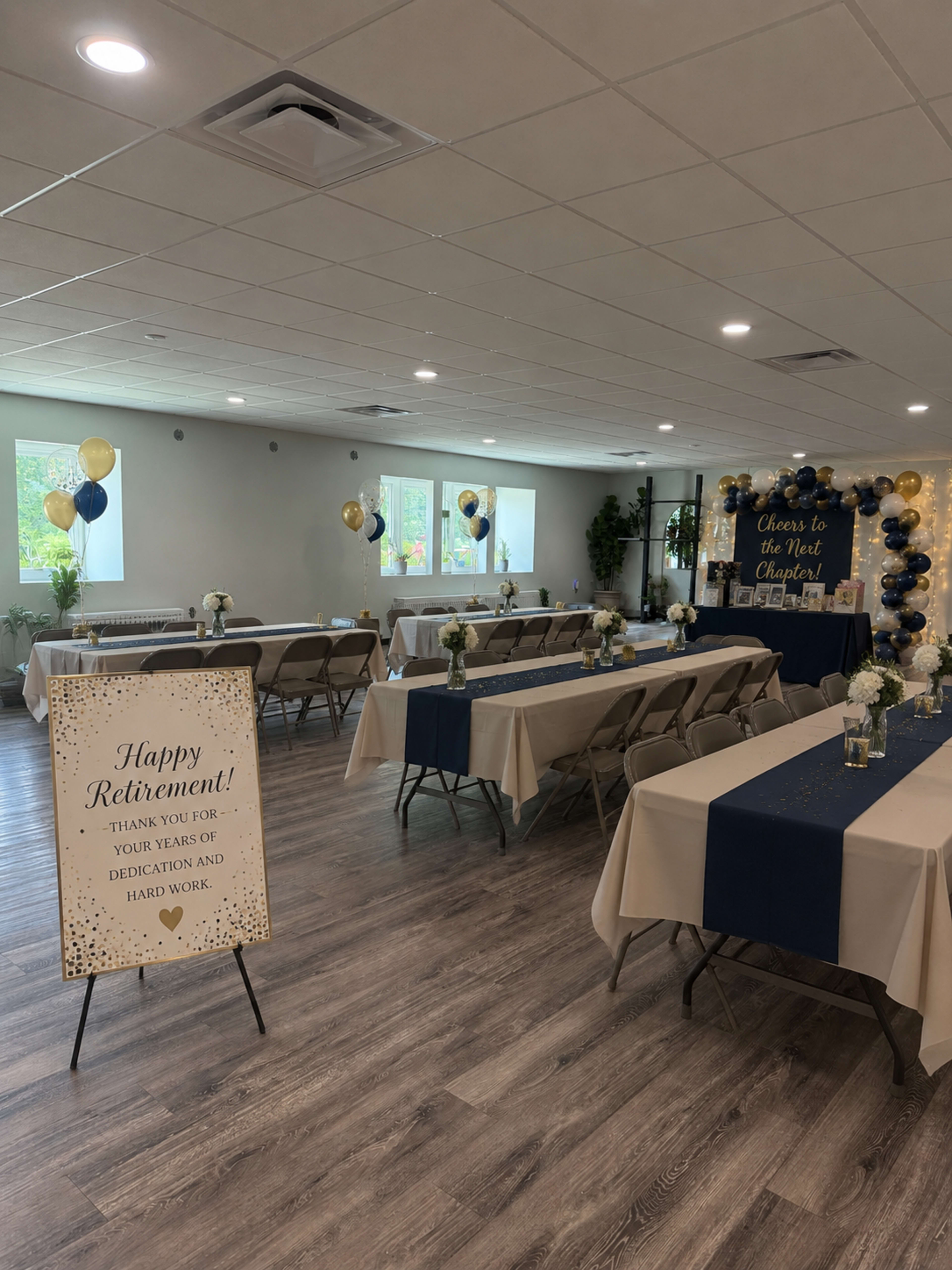 The image depicts a decorated event space set up for a retirement celebration, featuring tables with white tablecloths, gold and blue balloons, and a sign thanking the retiree for their years of service.
