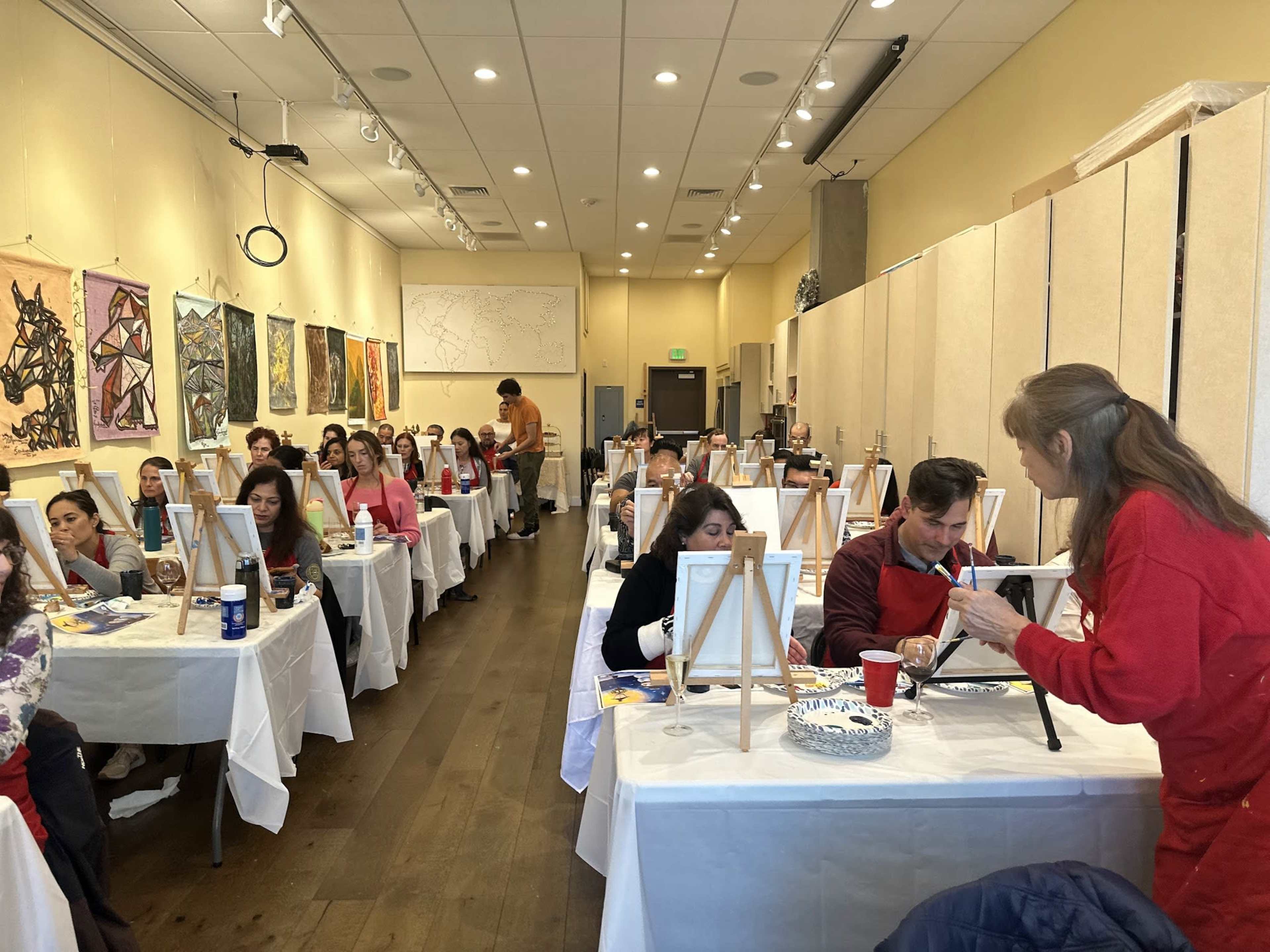 A painting class is in session with rows of participants seated at tables, each with a canvas and art supplies.
