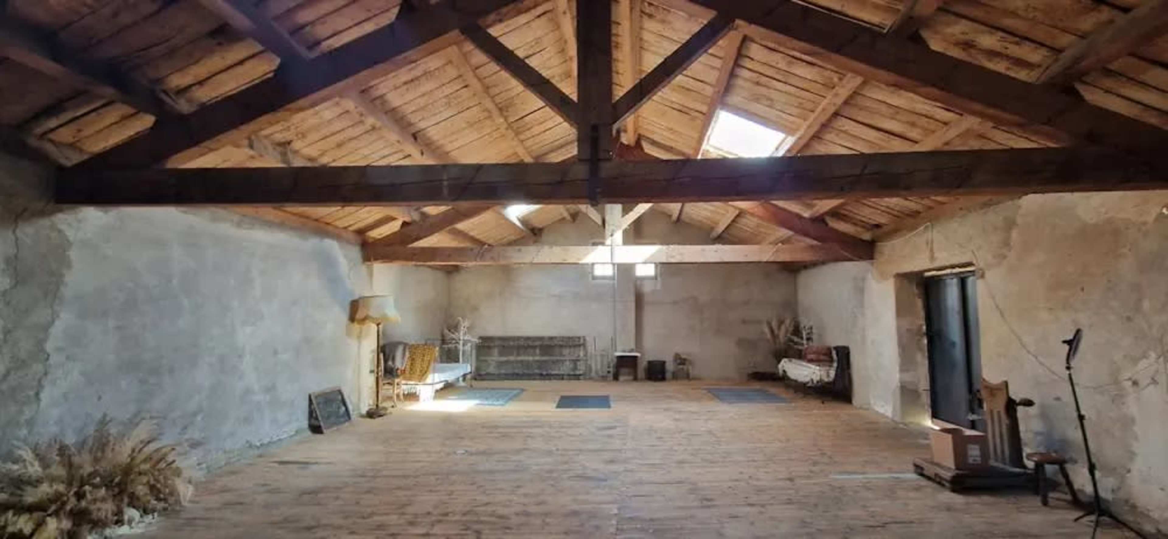 The image shows a spacious, interior room with a wooden beam ceiling, wooden floors, and sparse furnishings, including a sofa and a couple of benches along the walls.