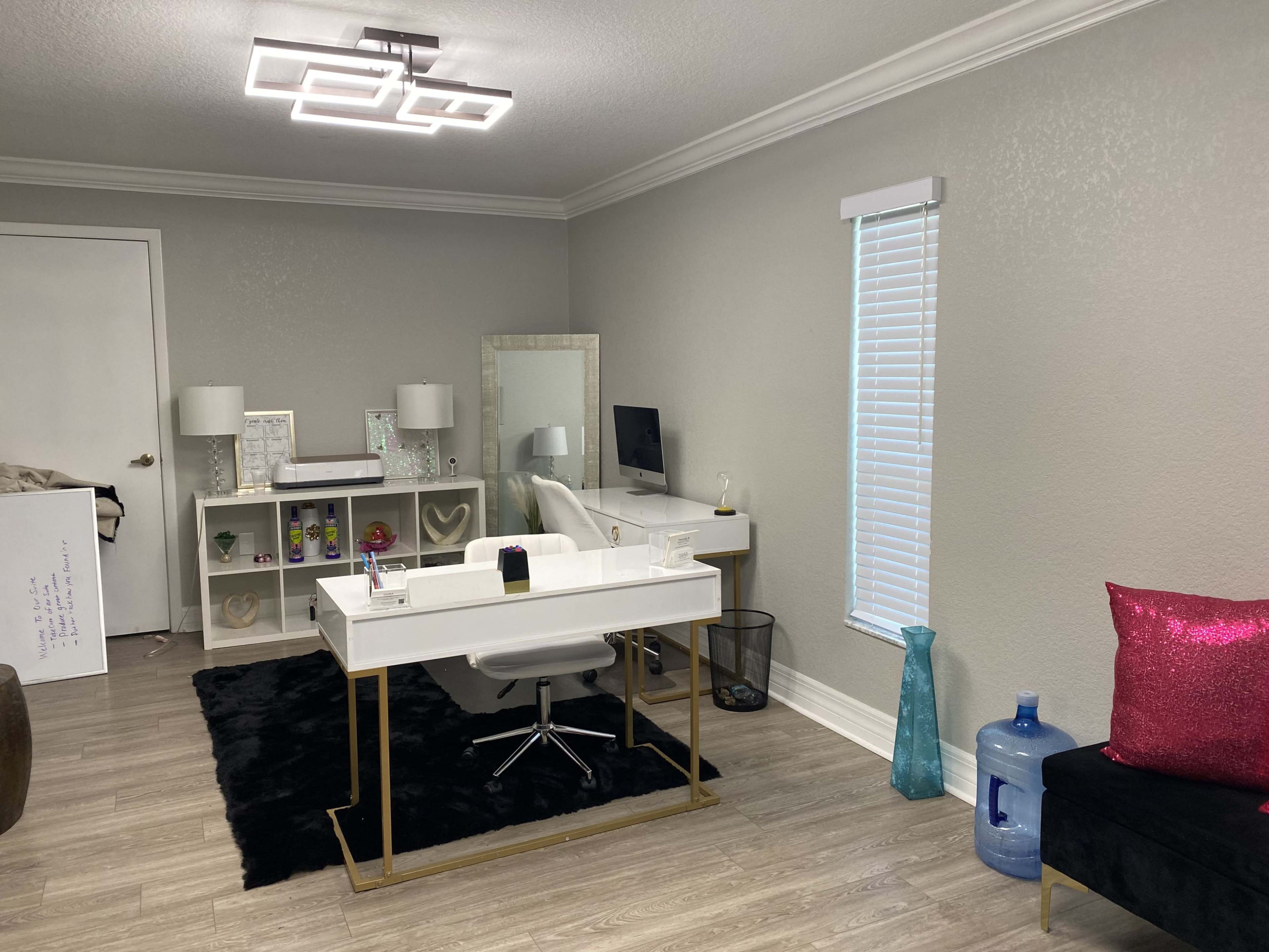 A modern office space features a white desk with a chair, a computer, decorative shelves, and various accessories against a light gray wall.