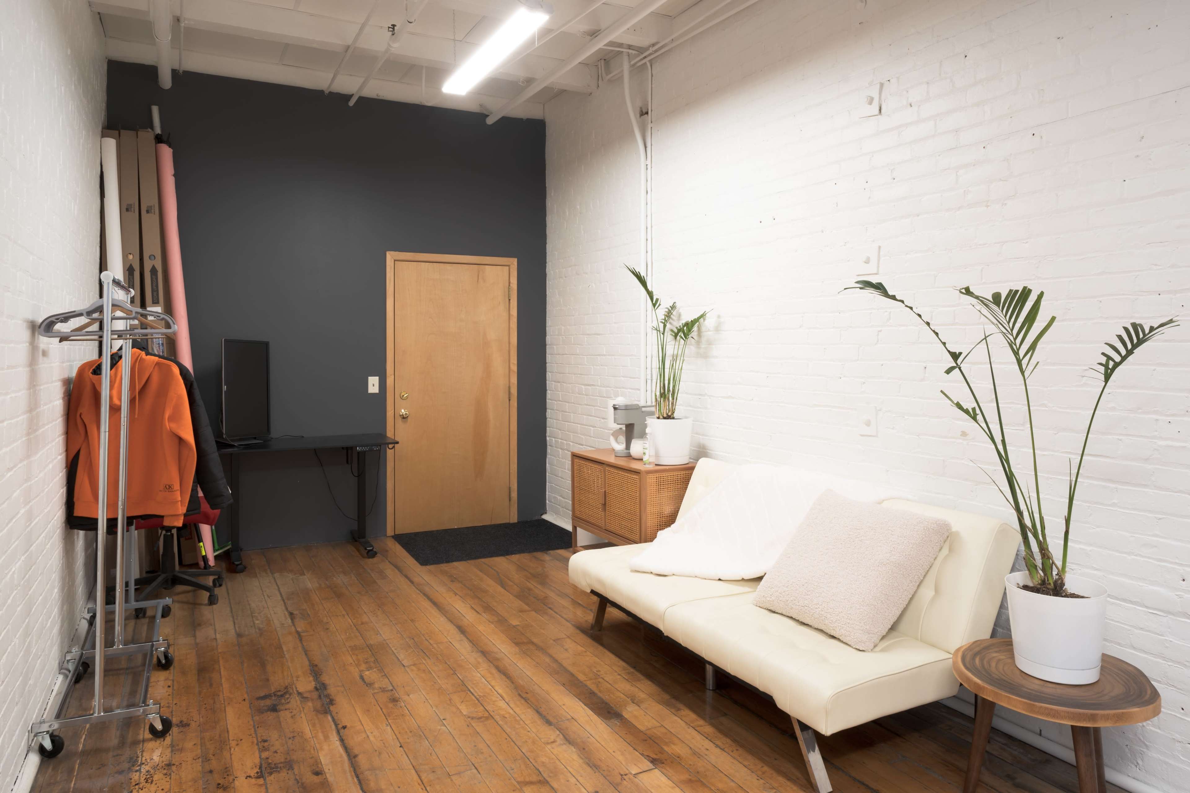 A small, well-lit room featuring a white brick wall, wooden flooring, a sofa, a door, a TV, and a plant.
