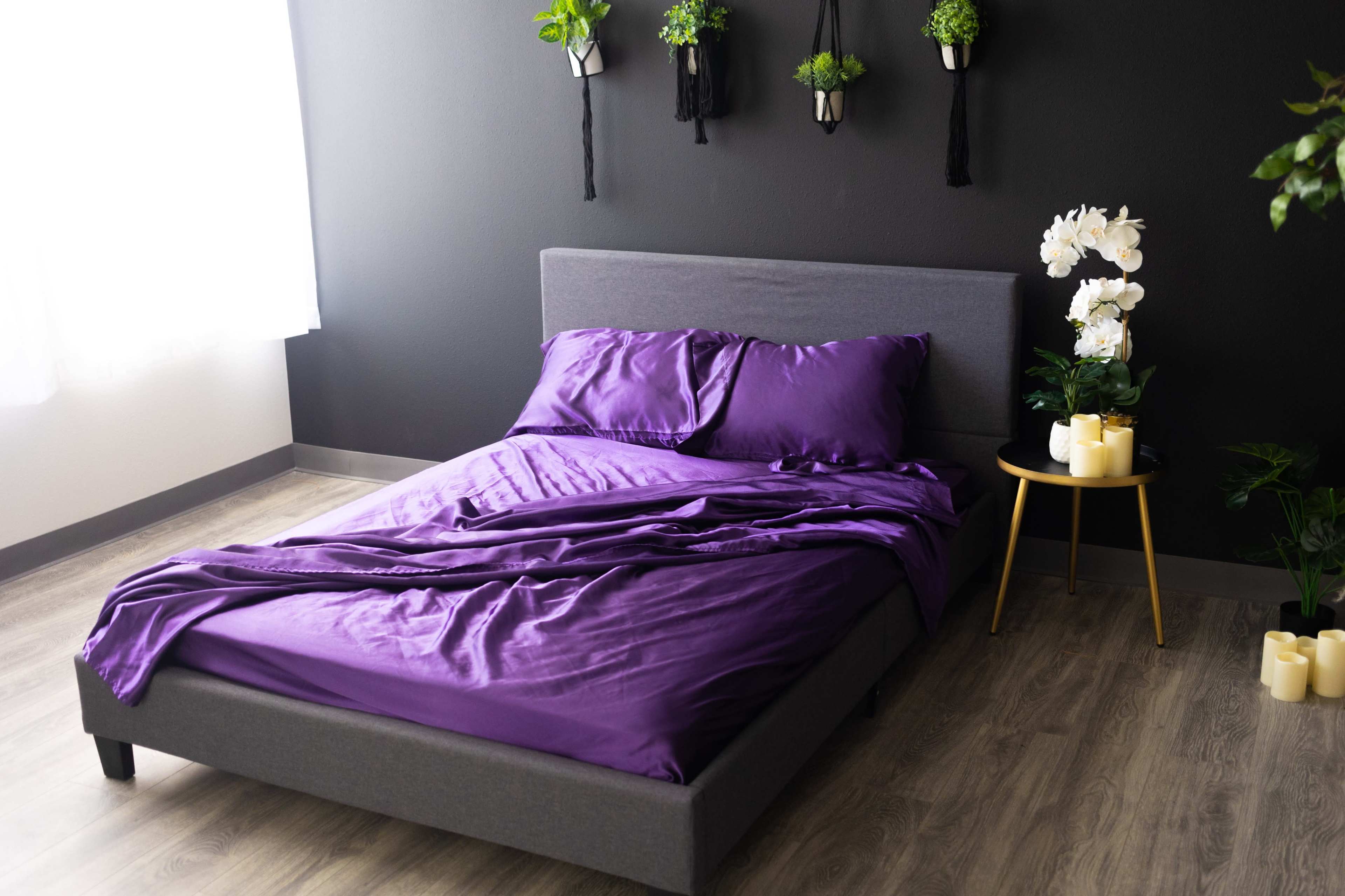 A gray bed with crumpled purple bedding is positioned next to a small gold table featuring candles and plants against a dark wall.