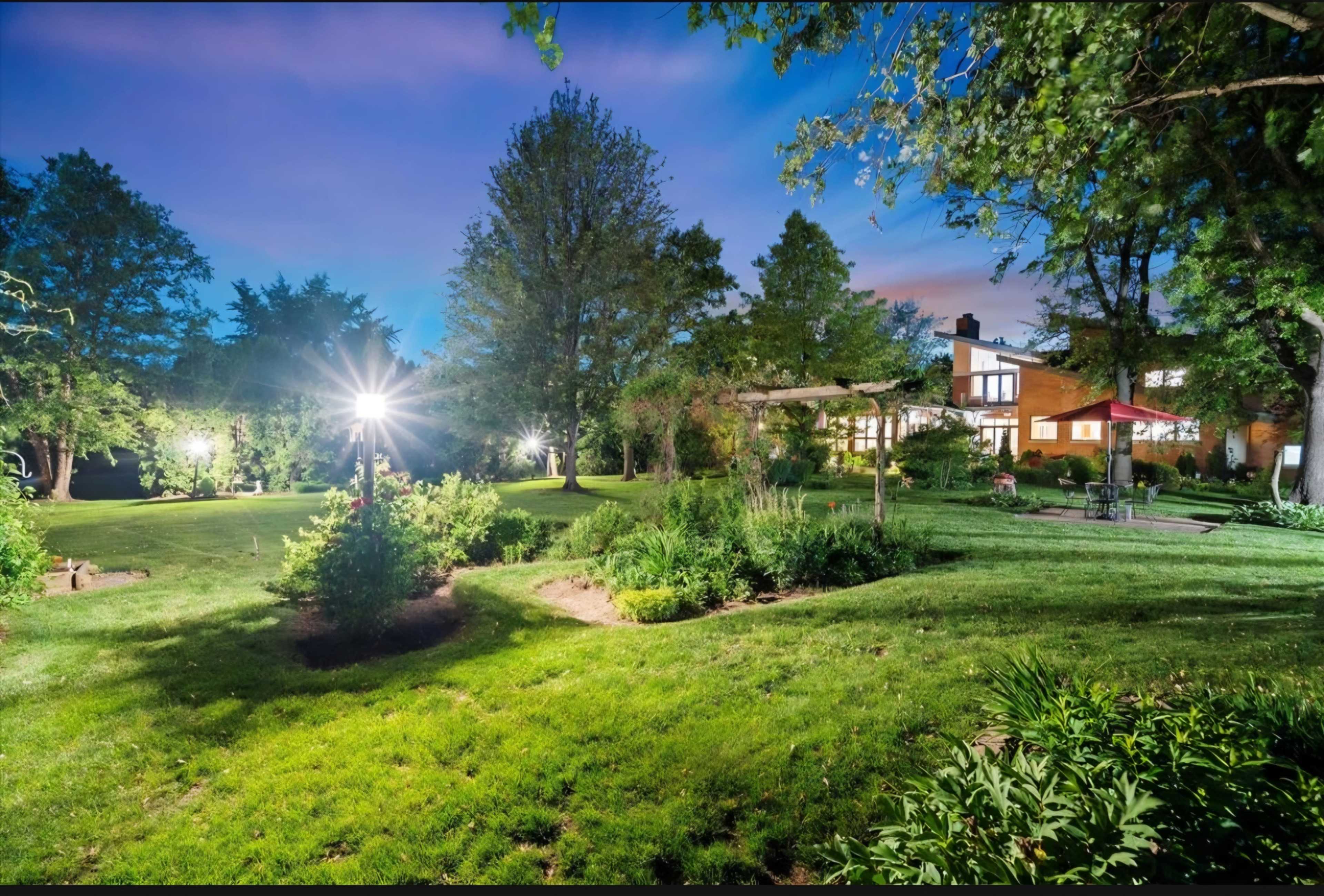 The image shows a well-maintained garden with lush green grass and various plants, illuminated by bright lights at dusk, with a house visible in the background.