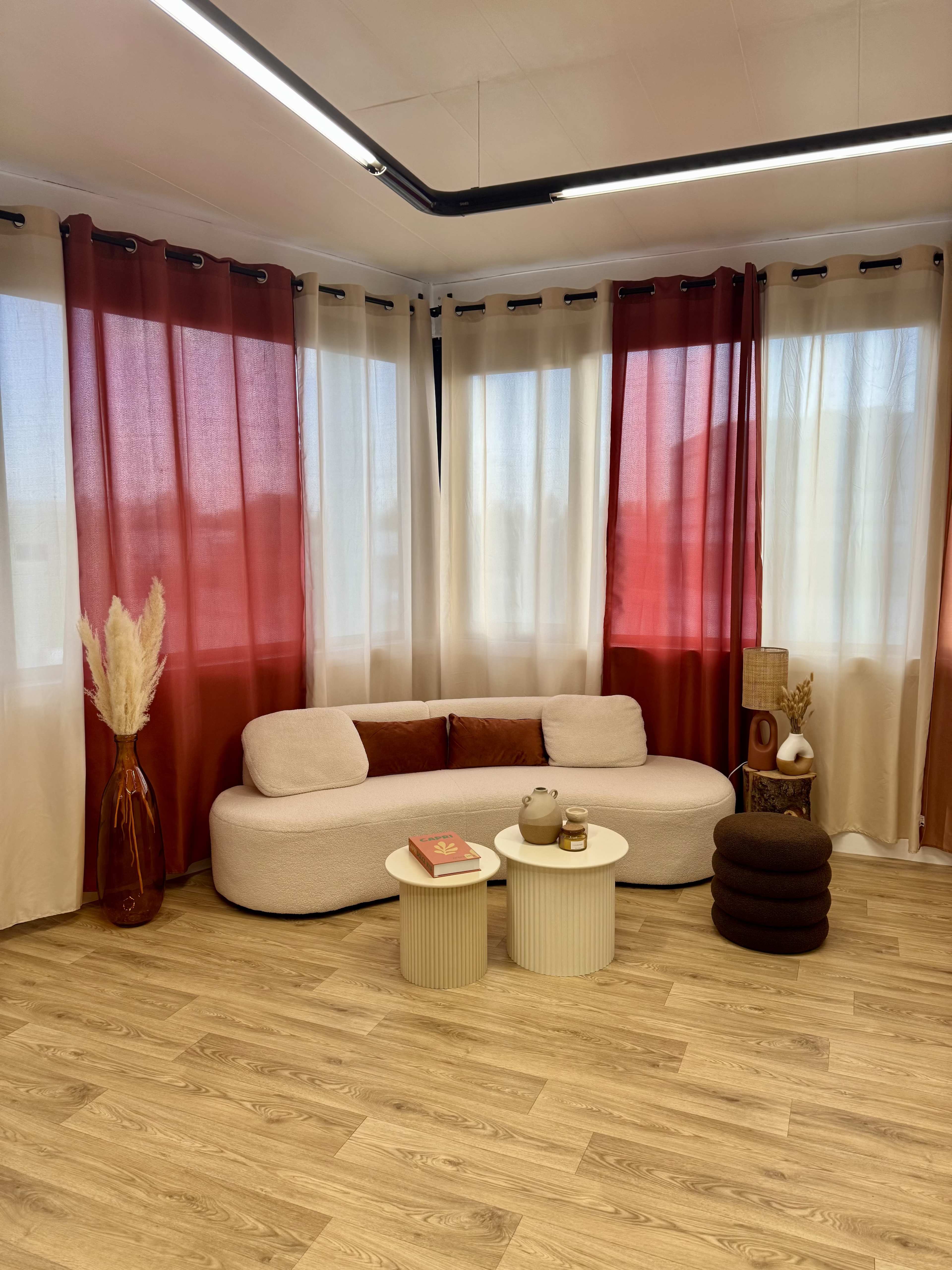 The image shows a cozy living area featuring a curved white sofa, two small round tables, and large windows dressed with red and cream curtains.