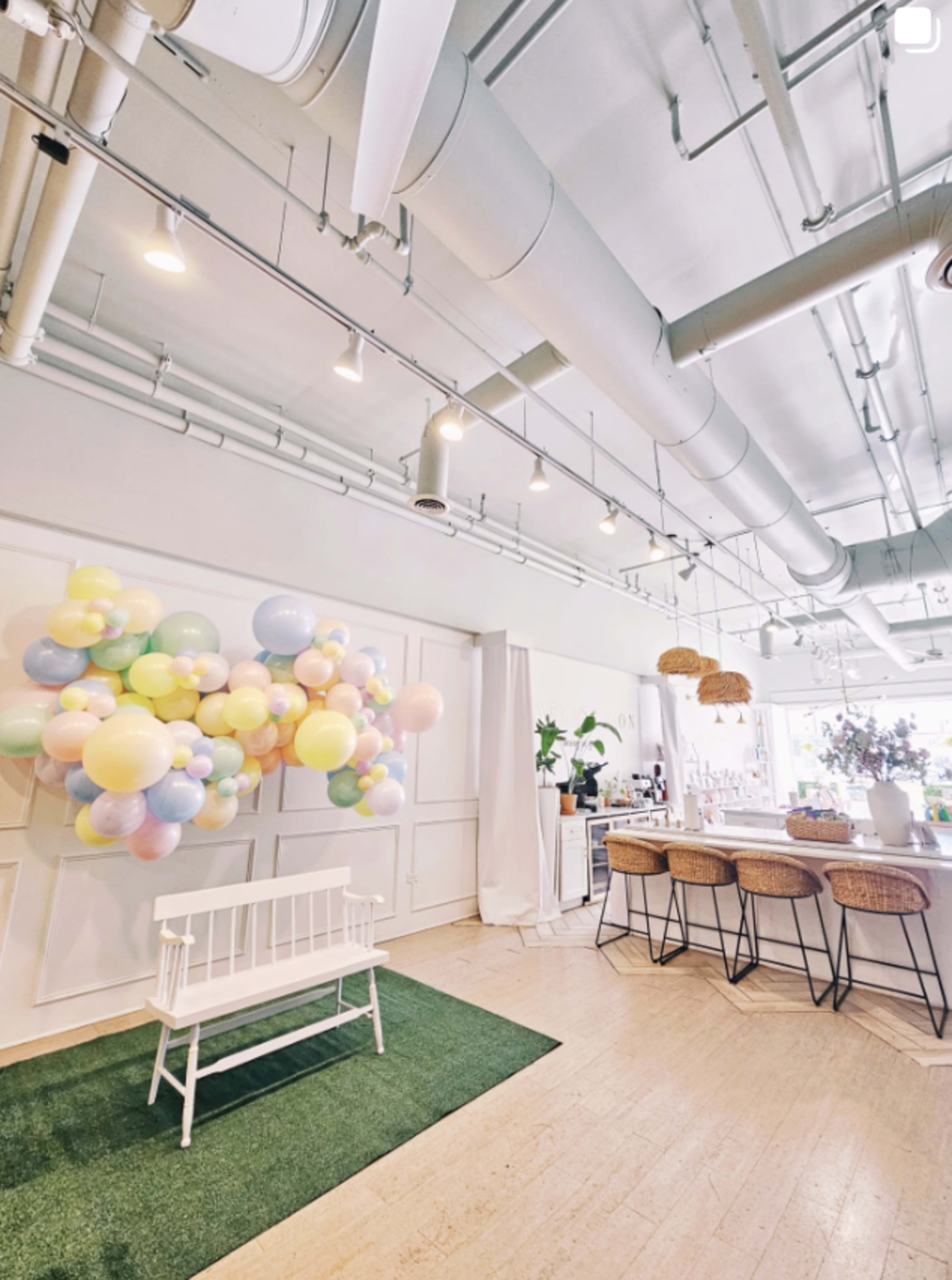 The image shows a bright, modern space with pastel-colored balloons on one wall, a white bench, and a bar with woven stools.
