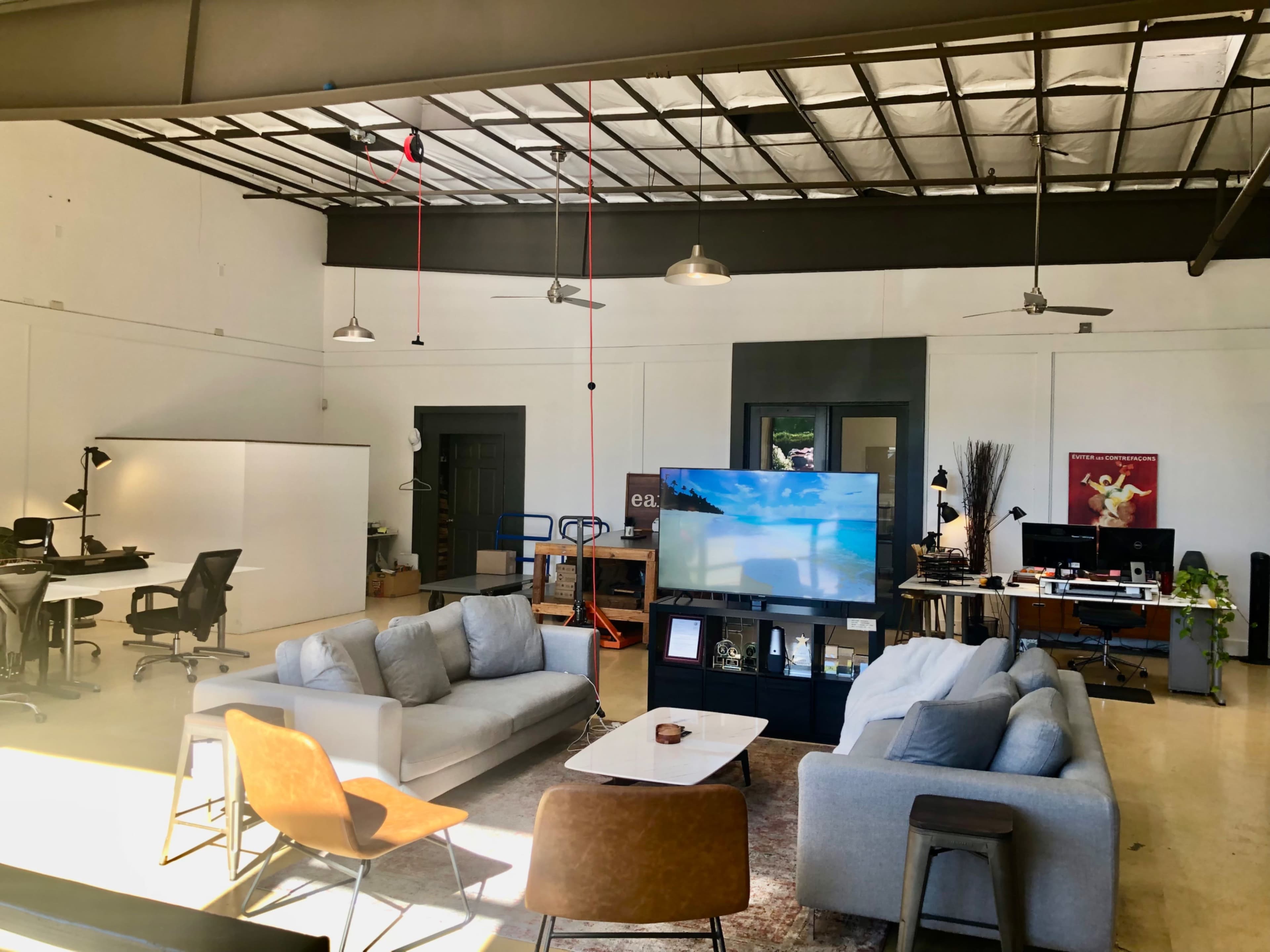 A spacious office interior with modern furniture, including two sofas and a television, surrounded by workstations and desk chairs.