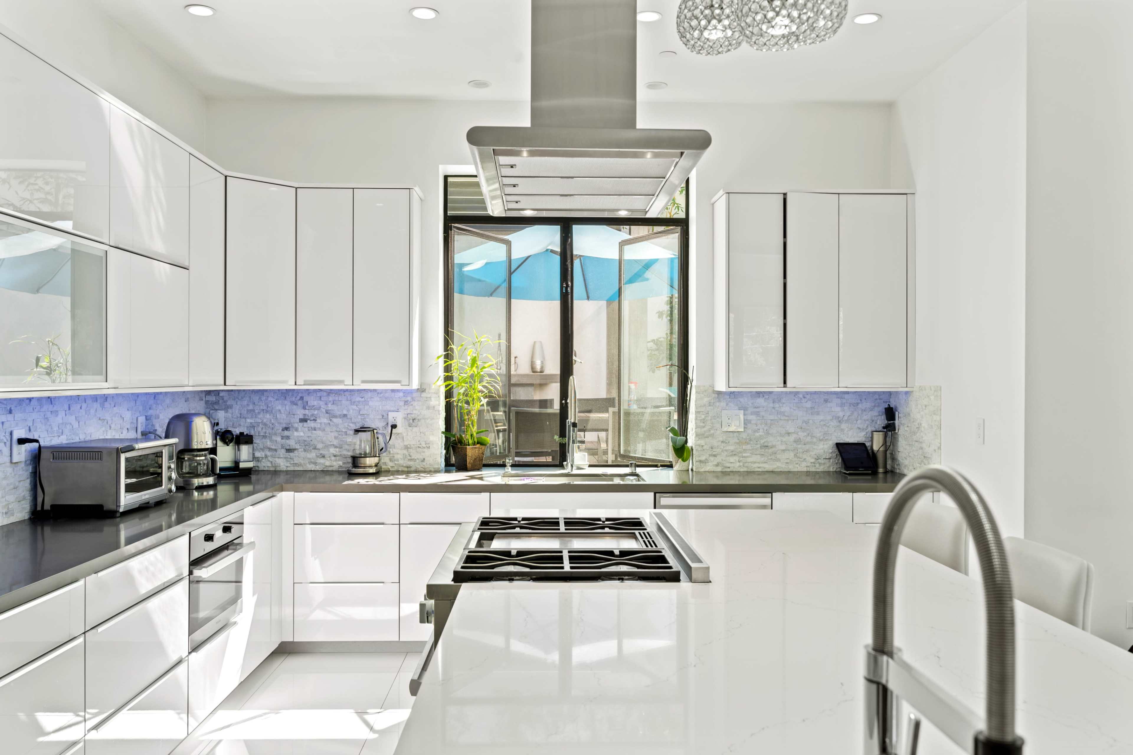 The kitchen features sleek white cabinetry, a central island with a stovetop, and a large window displaying blue patio umbrellas.