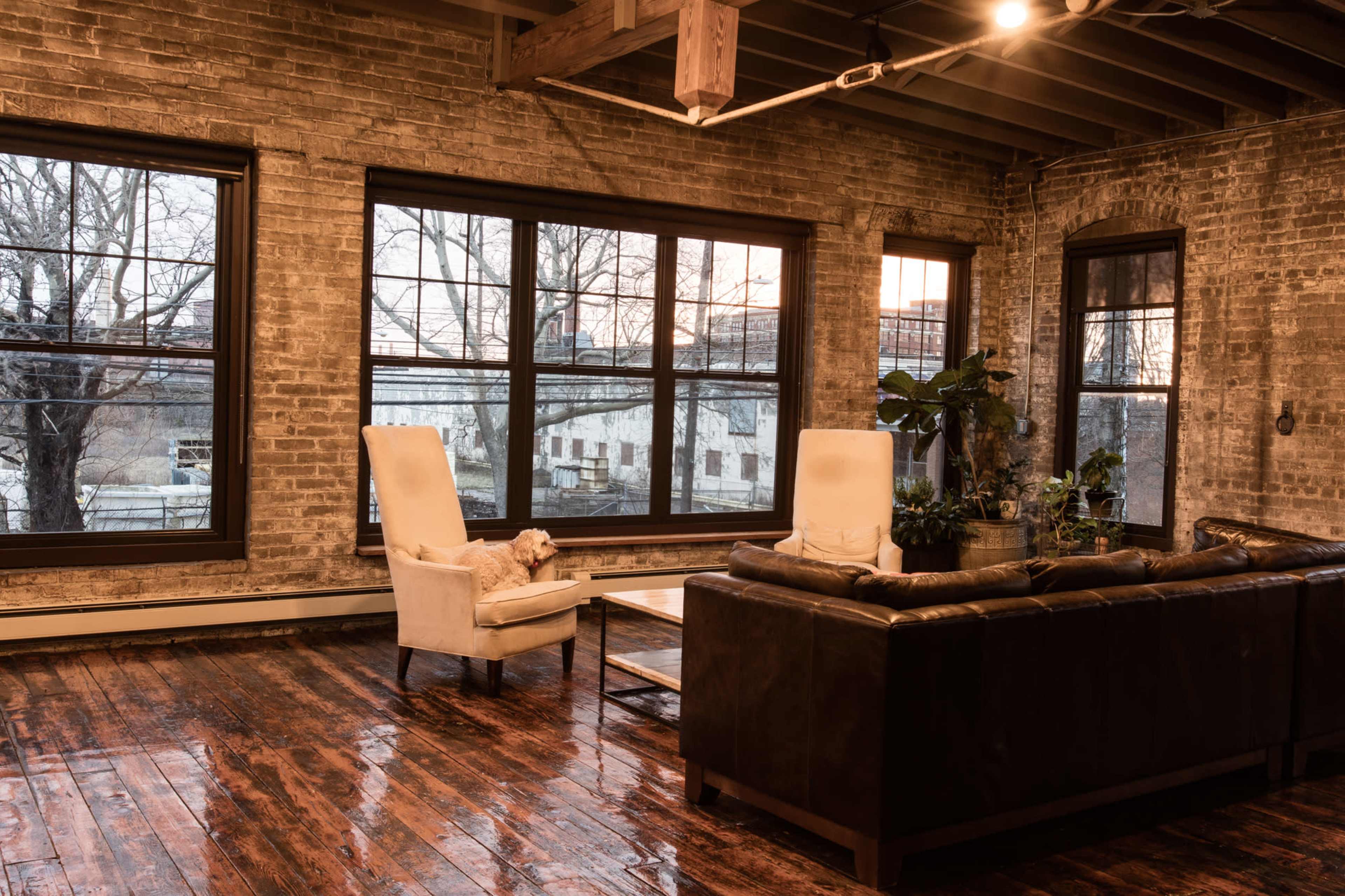 The image shows a modern living room with exposed brick walls, large windows, a leather sofa, a pair of armchairs, and wooden flooring.