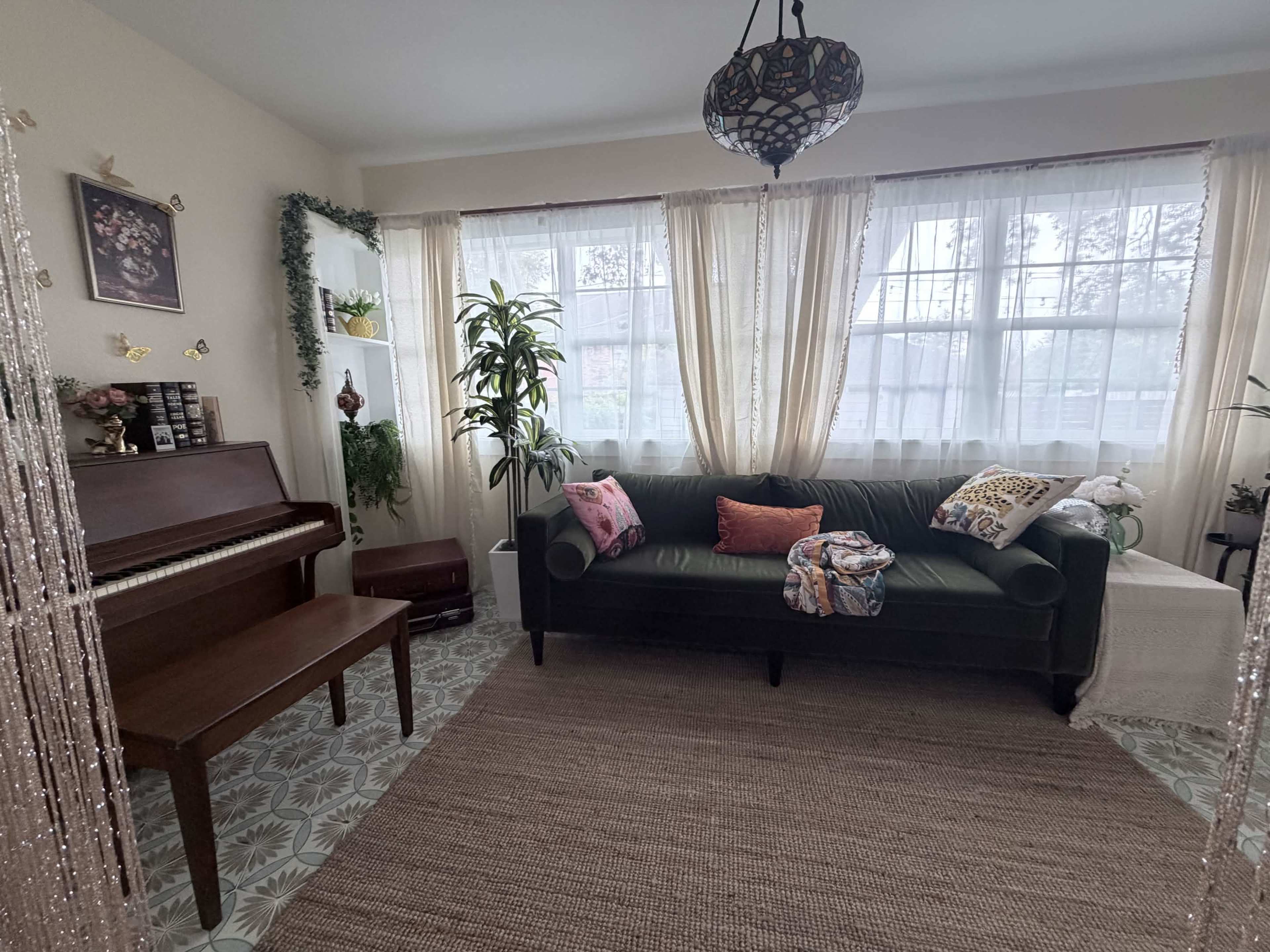 The image shows a bright living room with large windows, a dark green sofa, a piano, and various decorative plants.