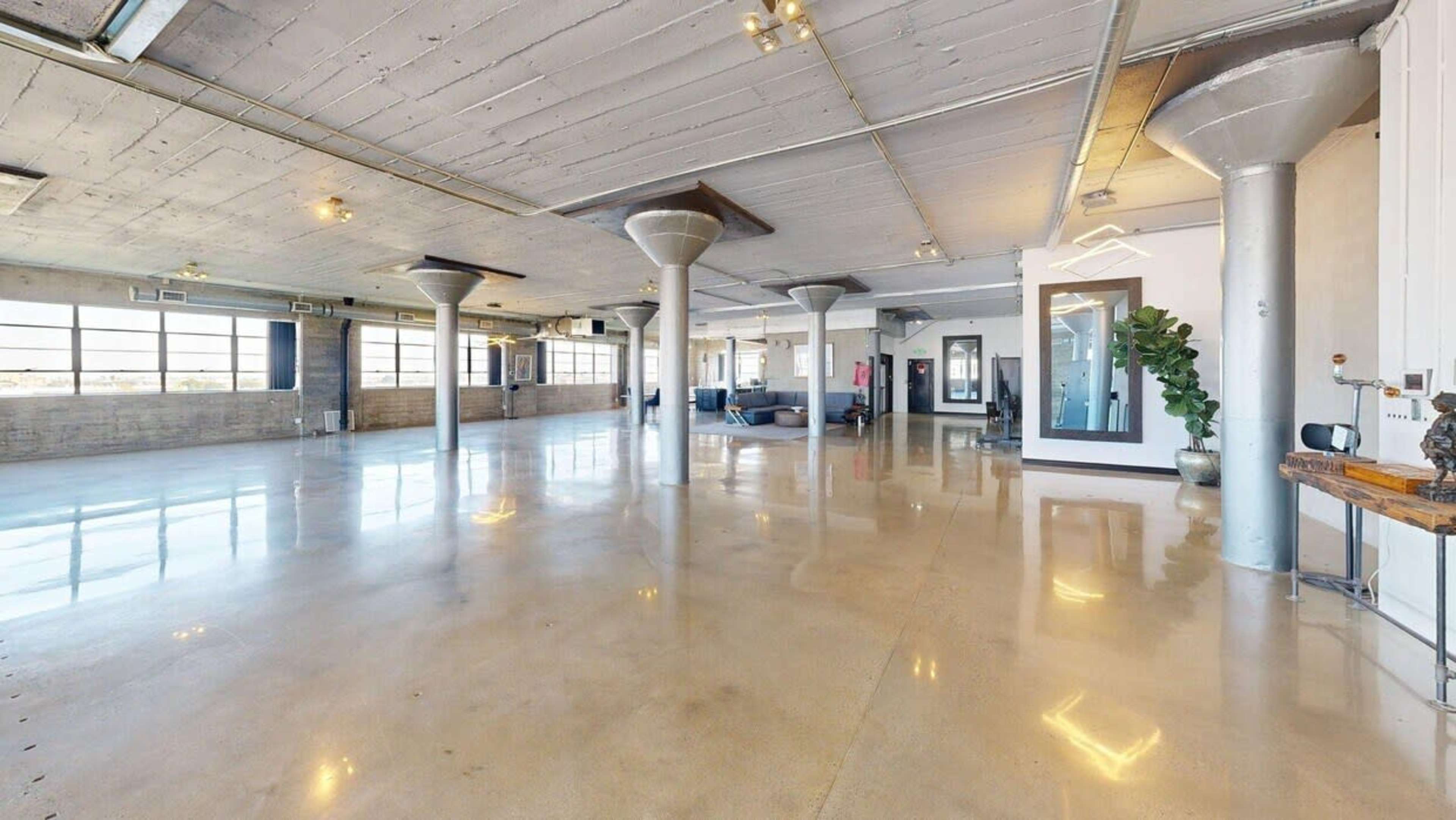 The image shows a spacious, industrial-style interior with polished concrete floors, large windows, and metal columns.