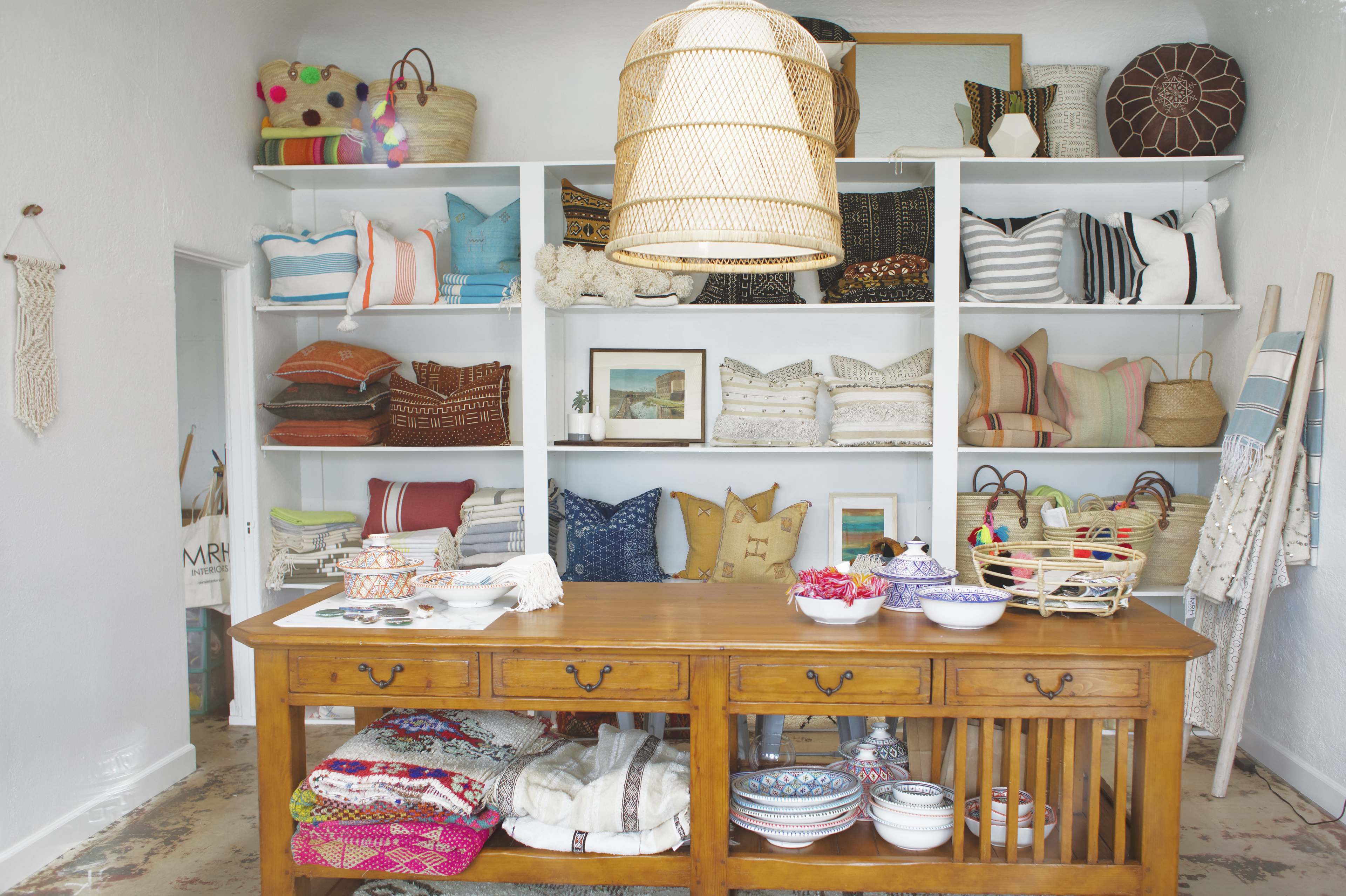 A well-organized retail space showcases various decorative pillows, woven baskets, and ceramic bowls on shelves and a central wooden table.
