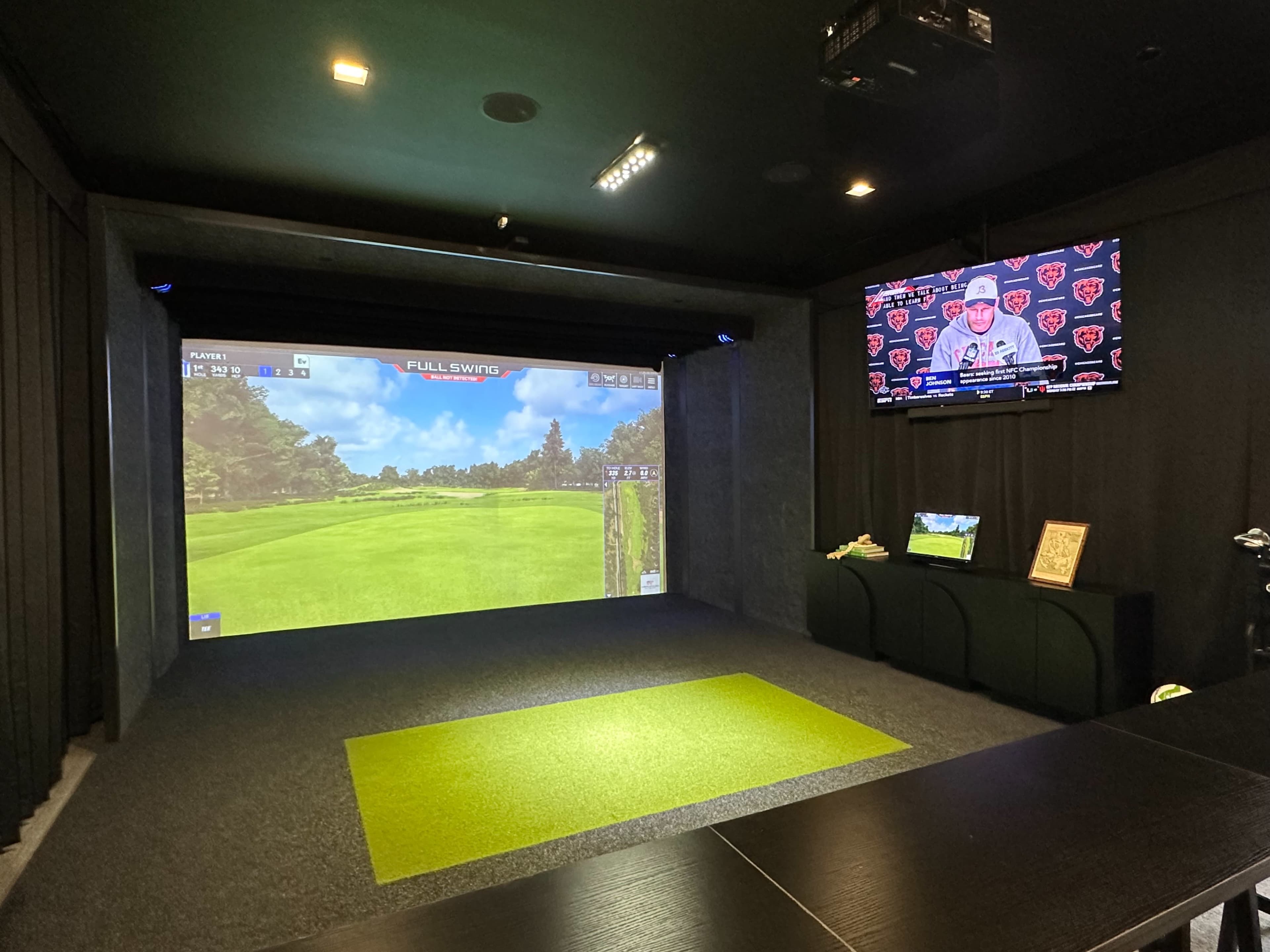 The image shows an indoor golf simulator with a large screen displaying a golf course and a TV mounted on the wall showing a sports press conference.