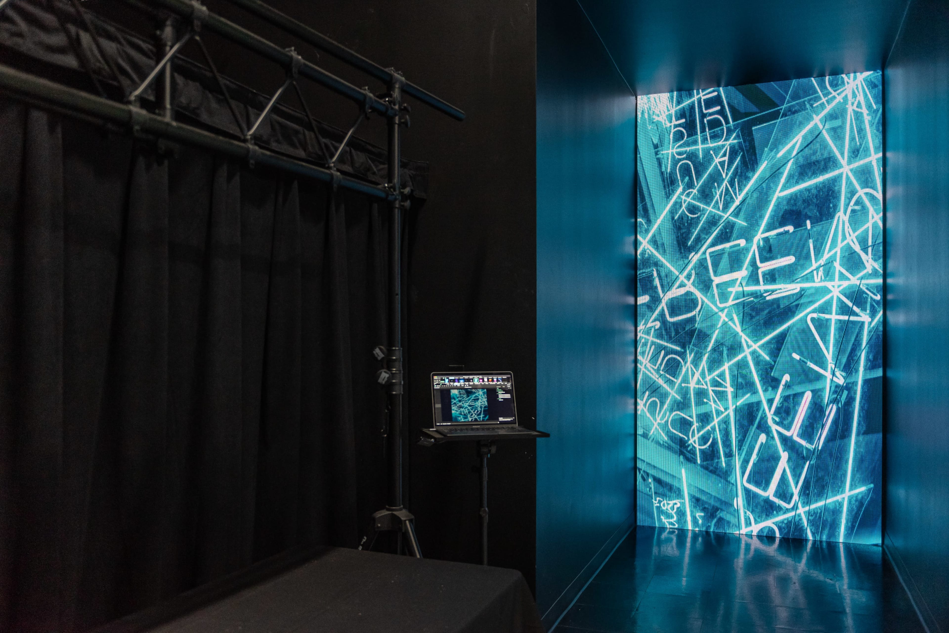 The image shows a darkened corridor leading to a brightly lit wall displaying blue digital text, with a black curtain and a laptop on a stand nearby.