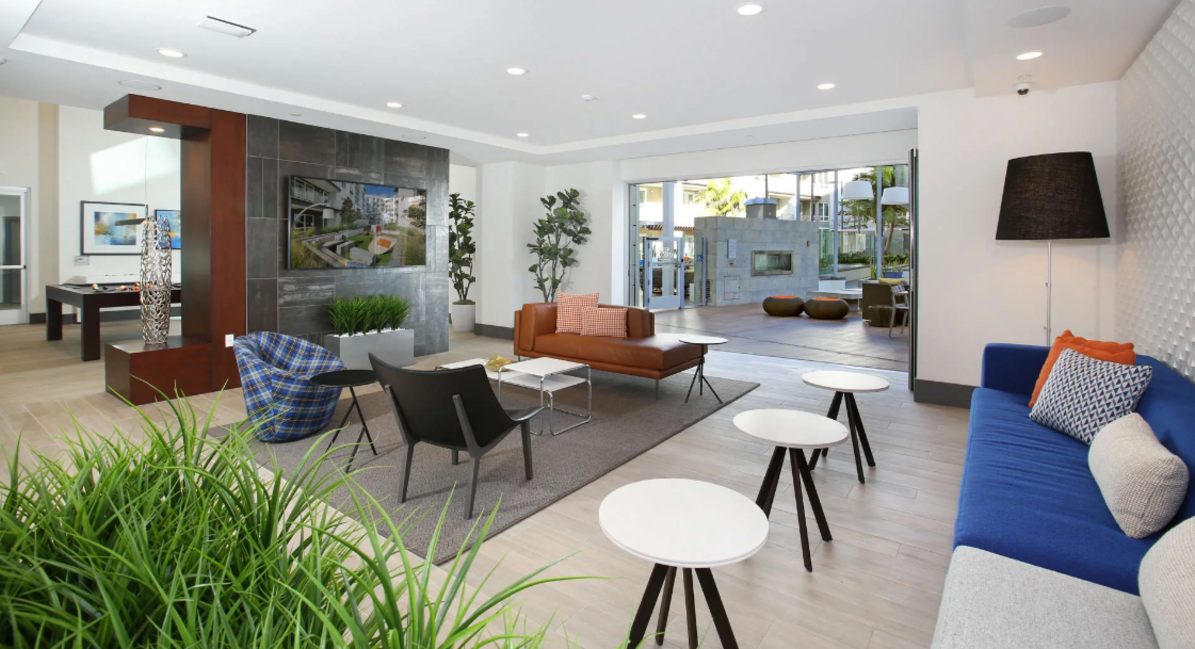 A modern lounge area featuring various seating options, a large flat-screen television on the wall, and large windows leading to an outdoor space.