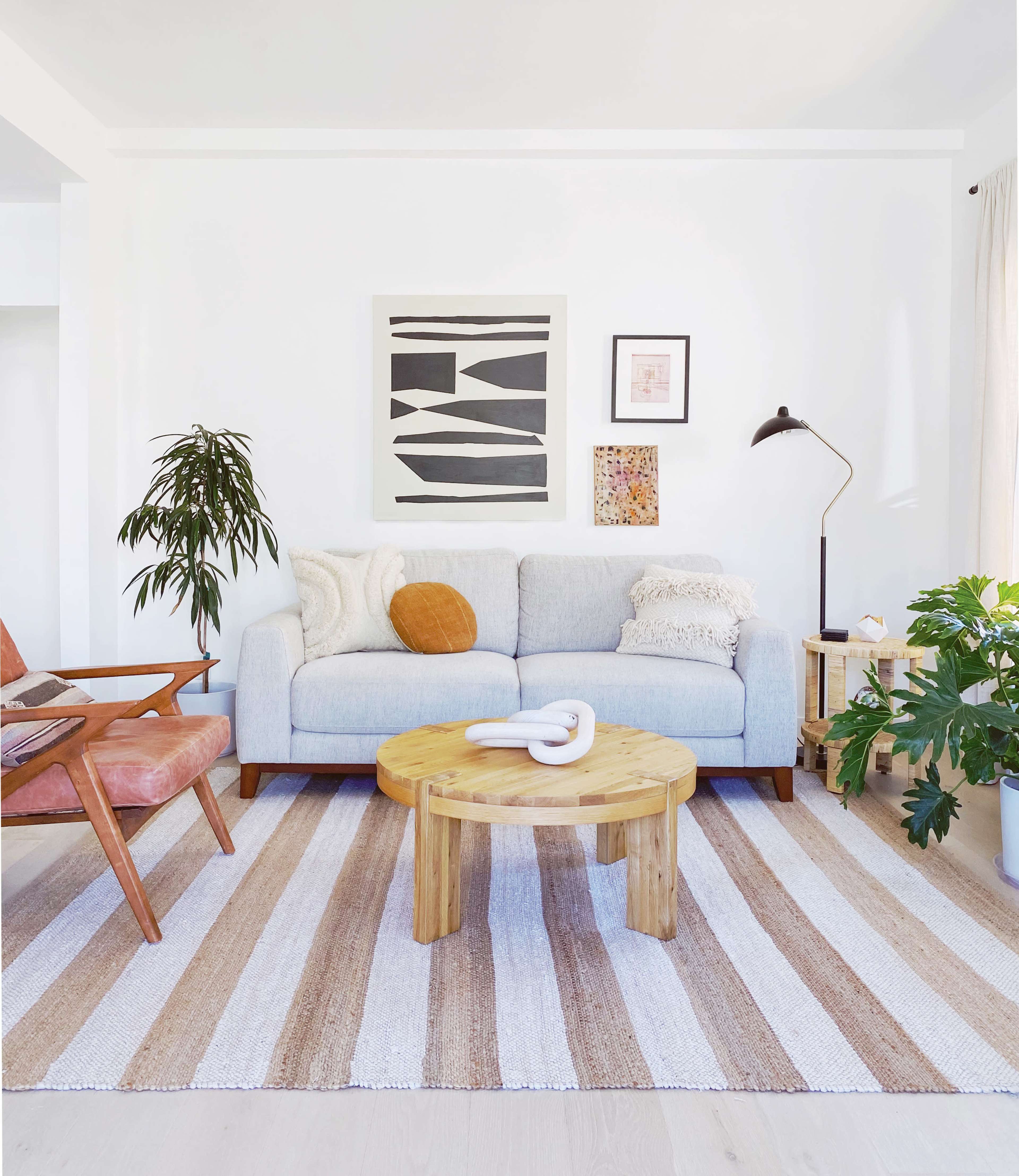 The image shows a modern living room with a light blue sofa, a round wooden coffee table, and a striped area rug, accompanied by plants and artwork on the walls.