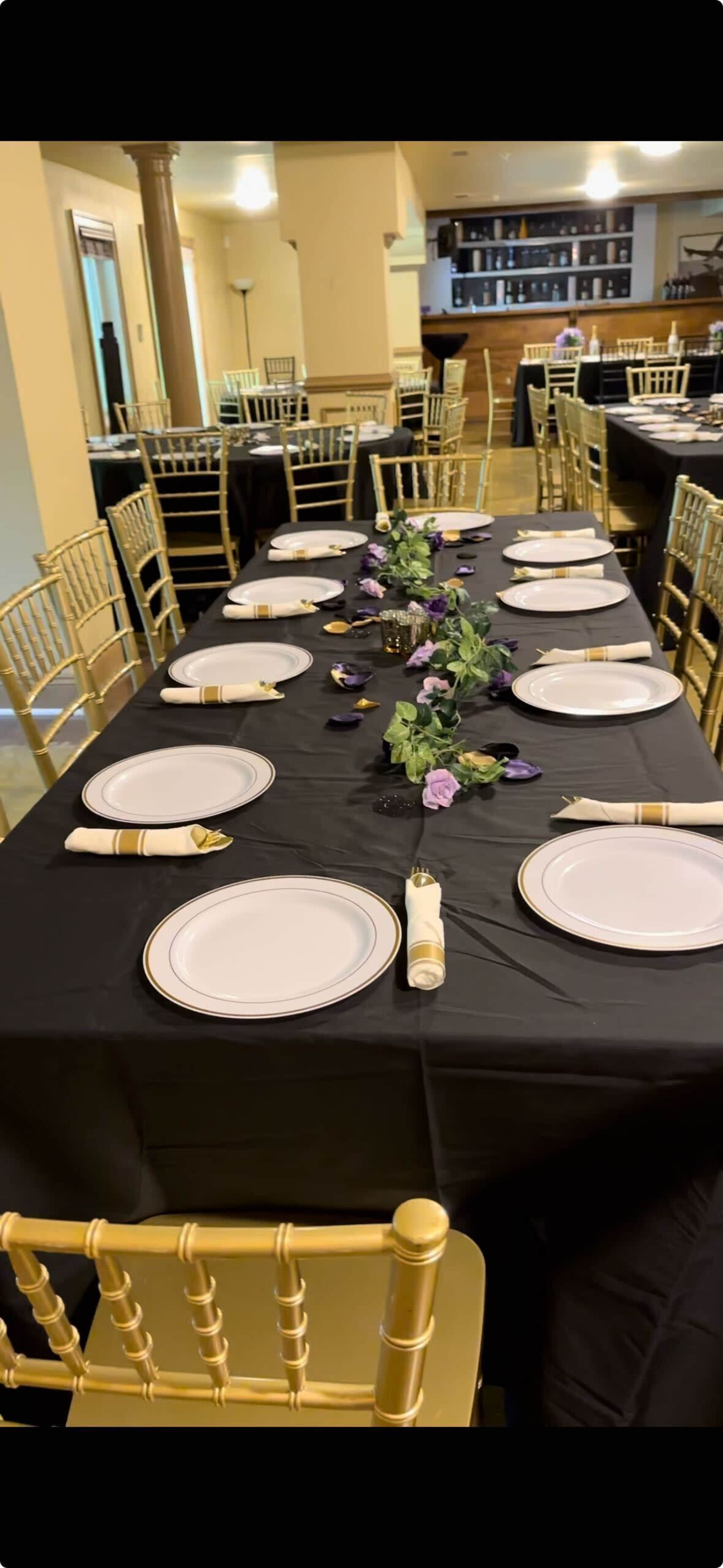 A dining setup features several tables arranged with gold chairs, black tablecloths, white plates, and floral decorations.