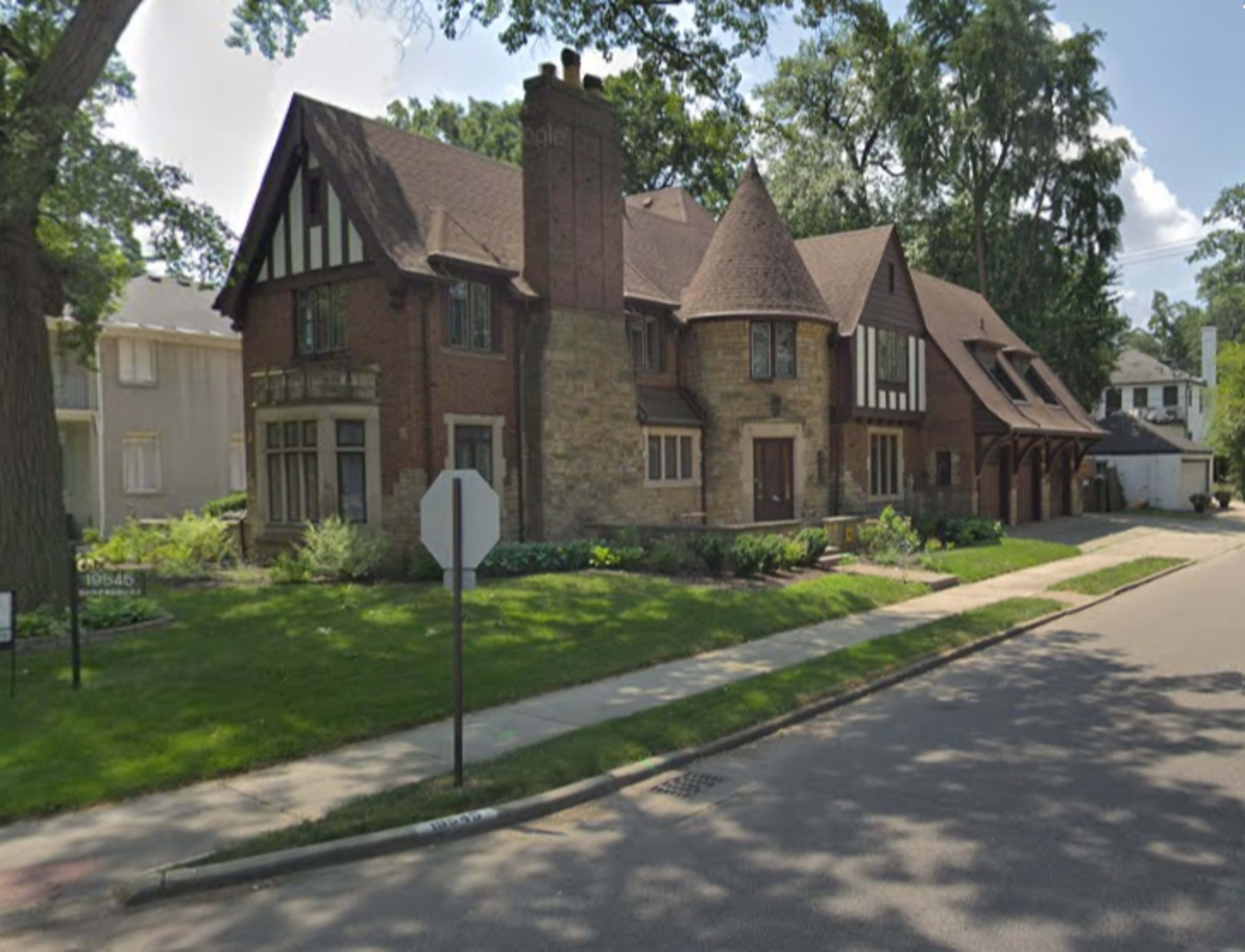 A large, multi-story house with a mix of brick and stone exterior features steeply pitched roofs and decorative elements, situated on a grassy corner lot.