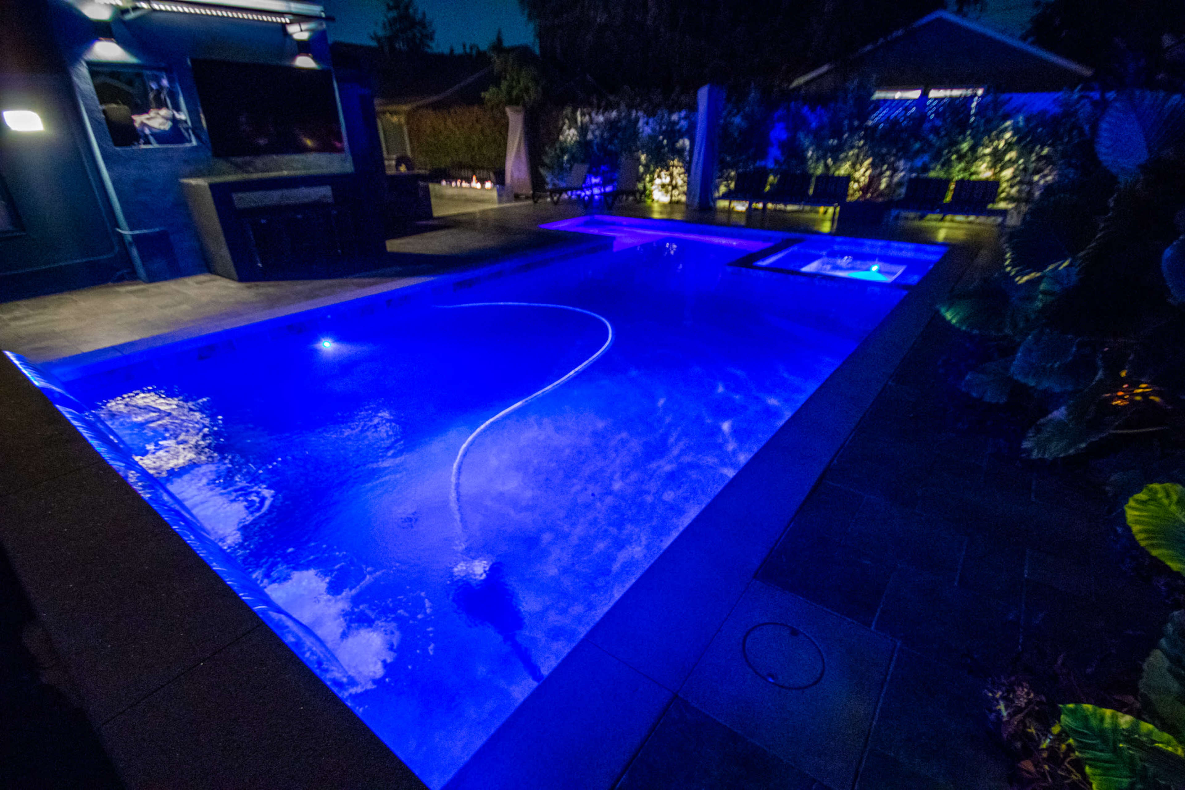 The image shows a modern pool illuminated with blue lights, featuring a spa area and surrounded by landscaped greenery.
