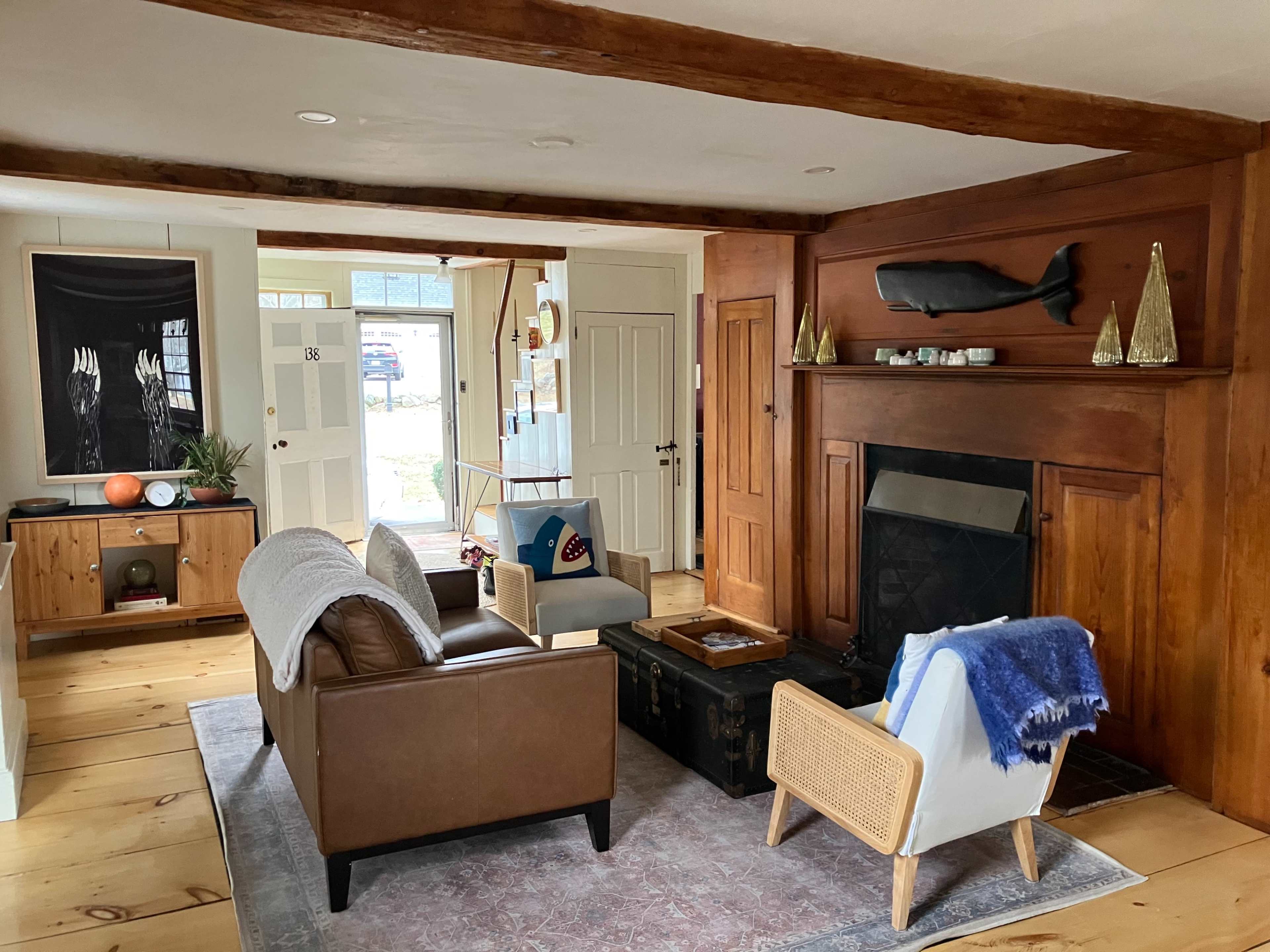 A cozy living room with a brown leather sofa, a blue chair, a wooden coffee table, and a fireplace, along with light-colored walls and wooden beams.