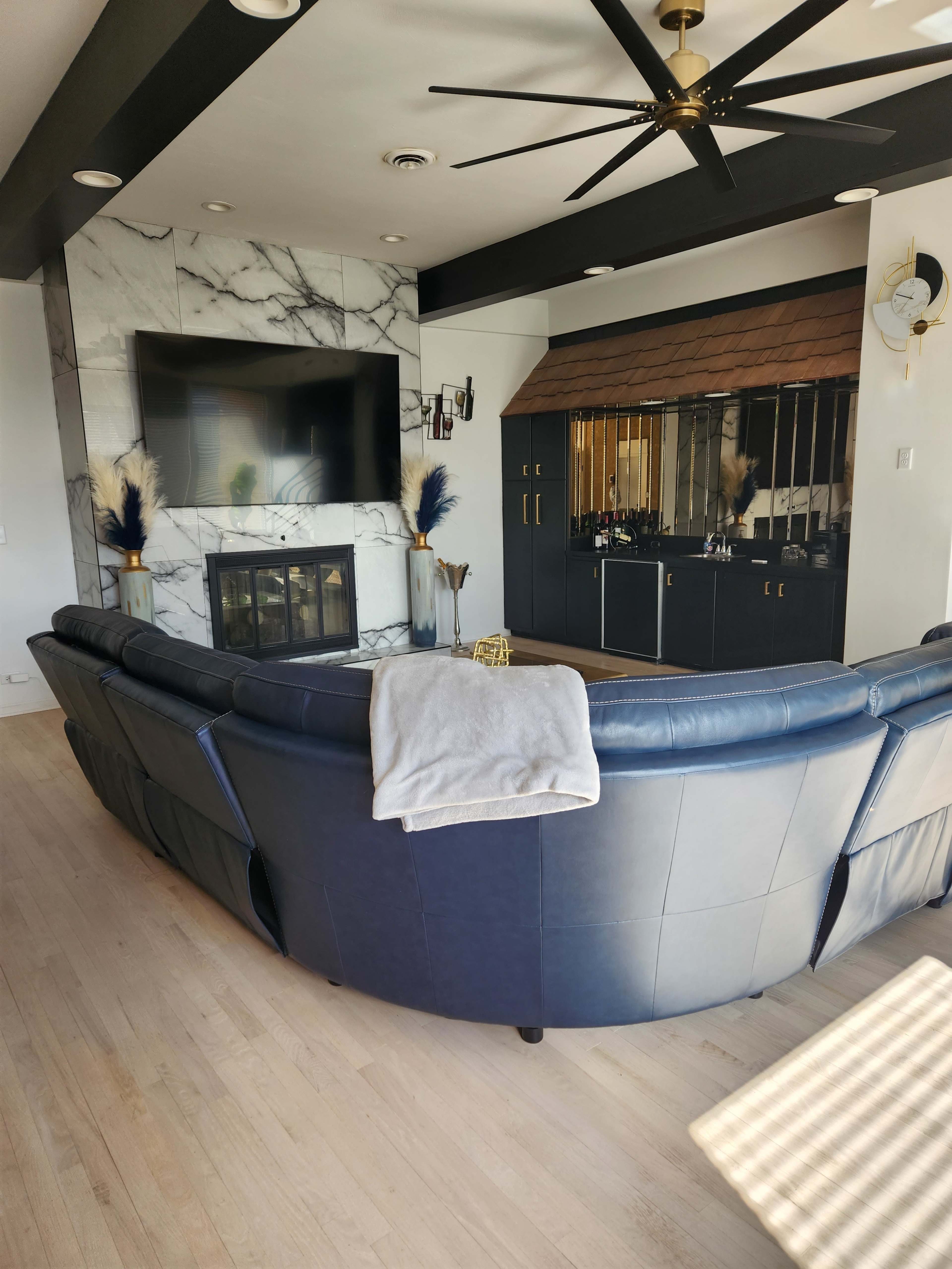 The image shows a modern living room featuring a large sectional sofa, a marble fireplace, and a dark cabinetry bar area with a large mirror.