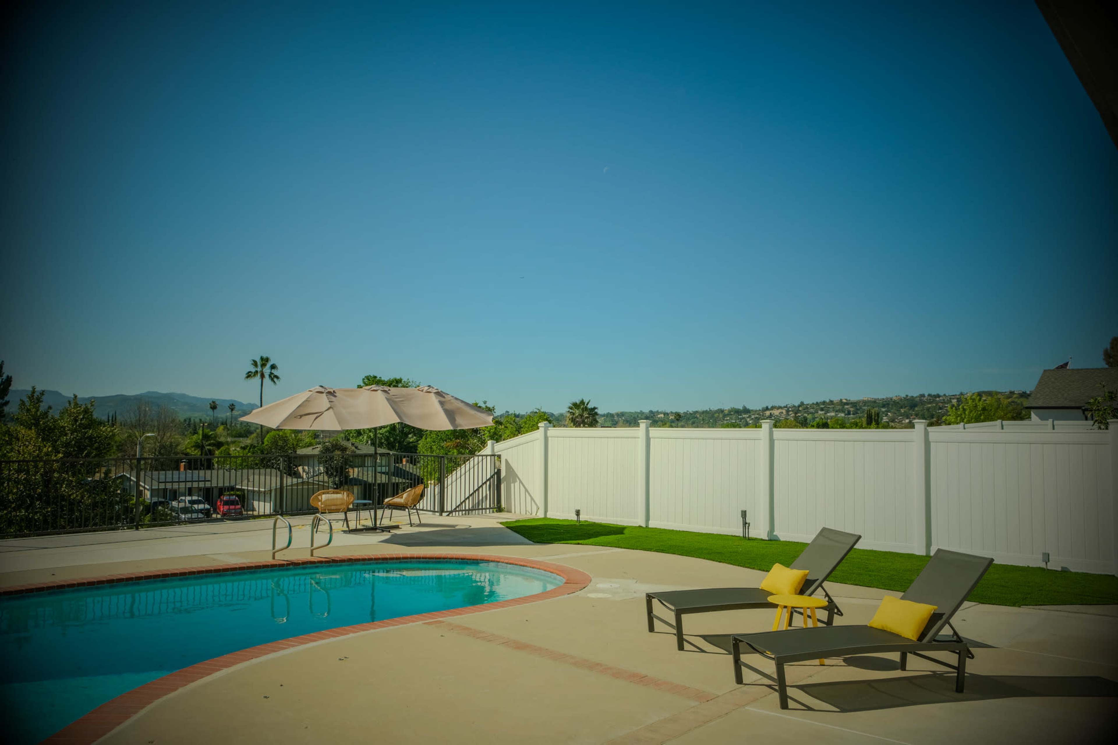 A swimming pool with two lounge chairs and an umbrella is set against a clear blue sky and a white fence.