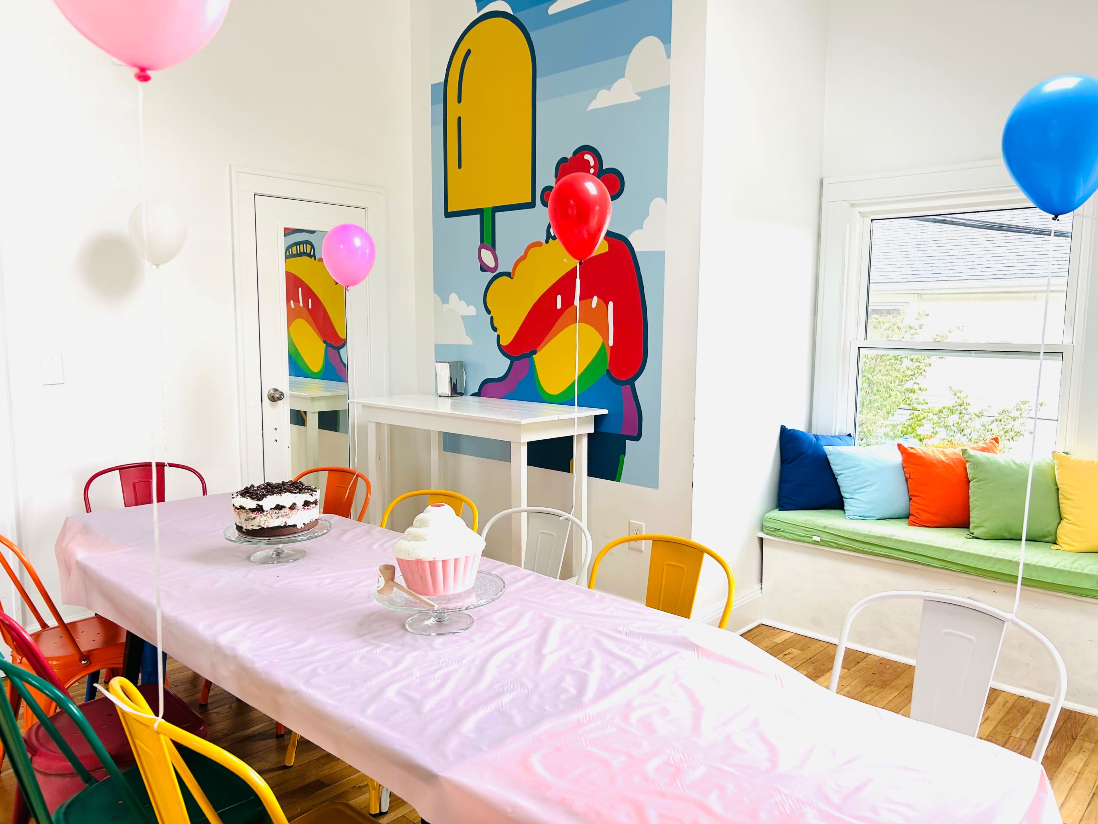 A long table covered with a pink tablecloth is set in a brightly colored room decorated with balloons and a mural featuring a cartoon character.