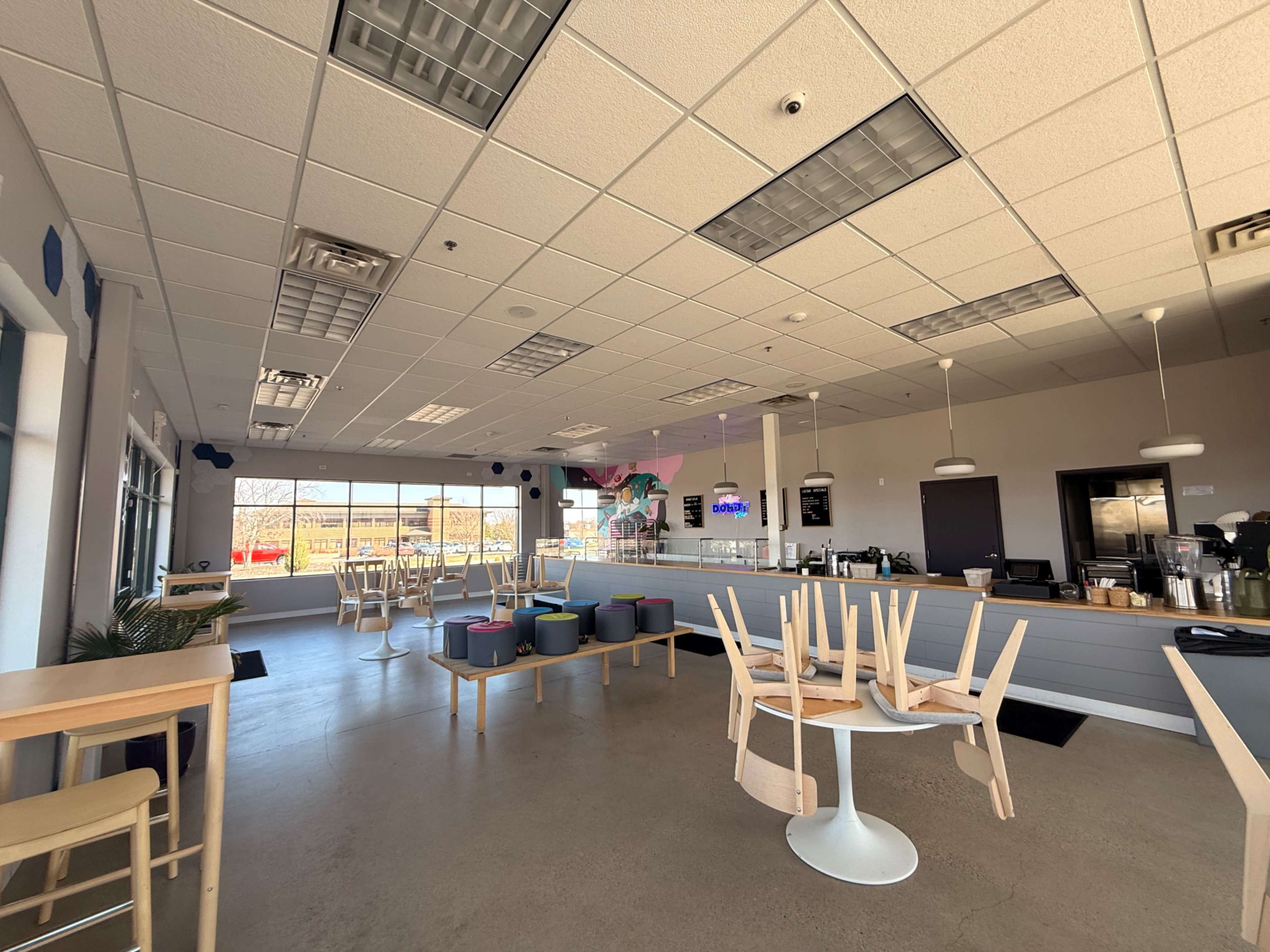 The image shows an empty café with tables and chairs stacked upside down and a large window offering a view outside.