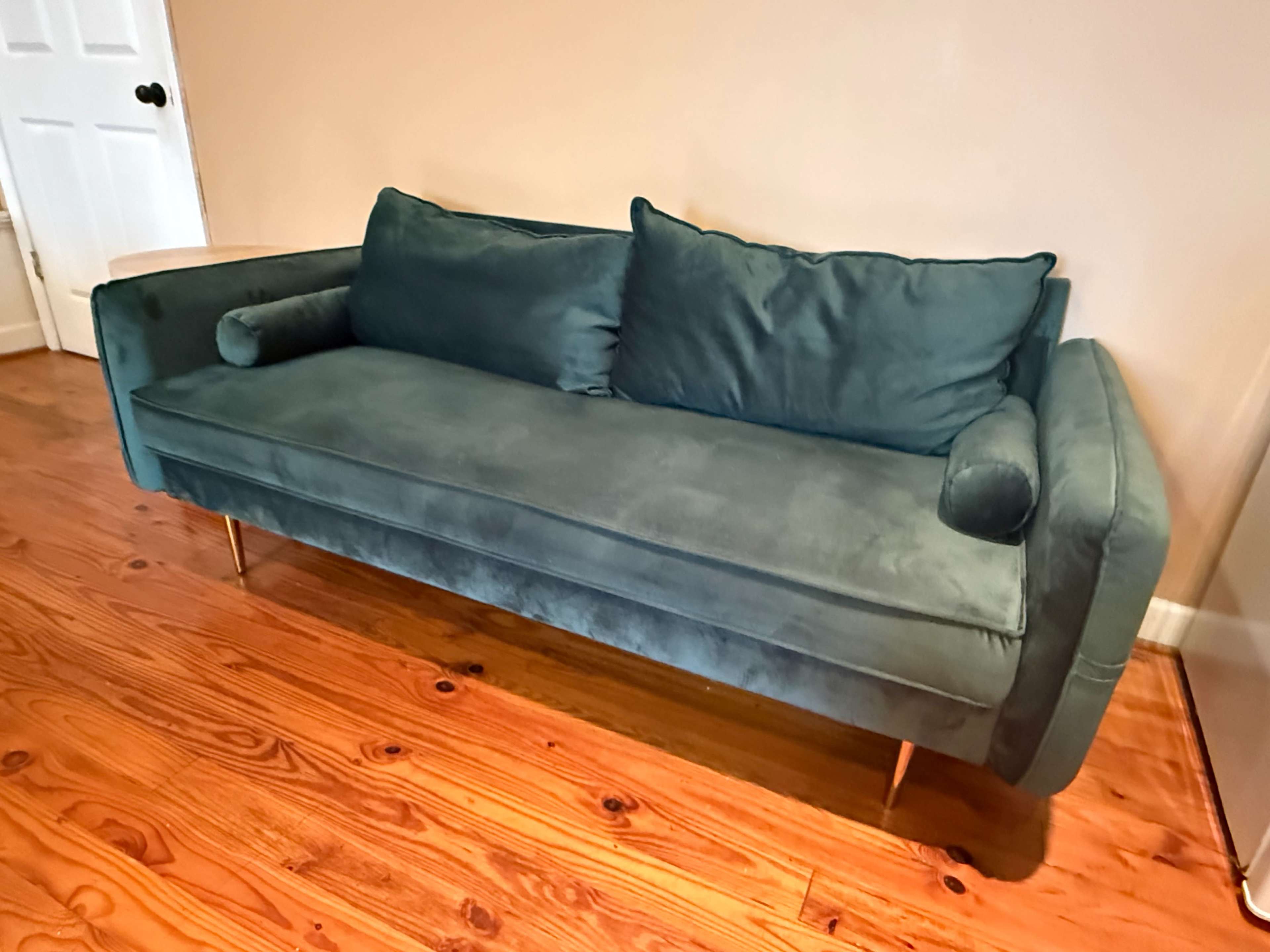 A green velvet sofa with two cushions rests on a wooden floor in a well-lit room.