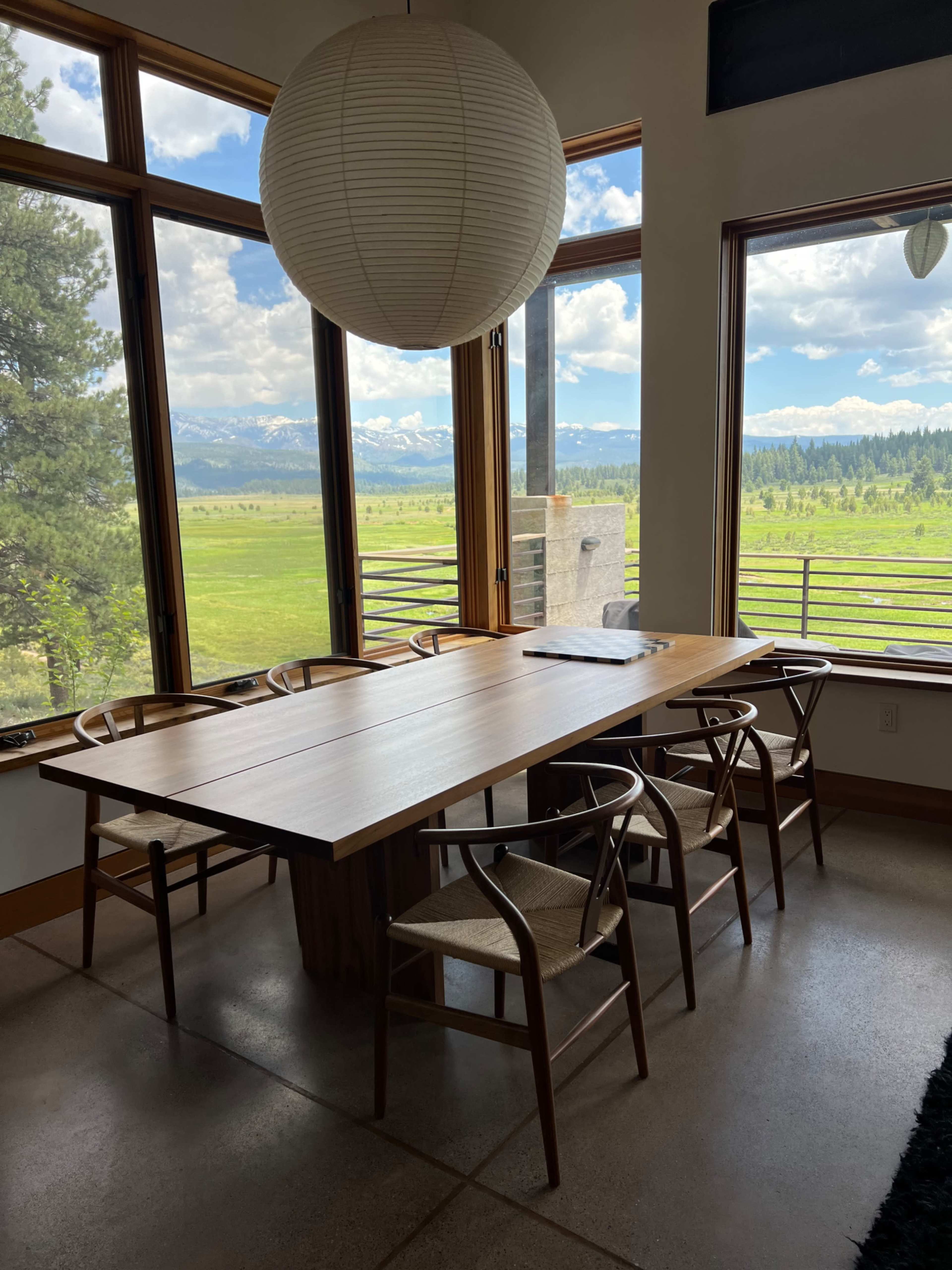 A wooden dining table is surrounded by six chairs in a room with large windows overlooking a green landscape and mountains.