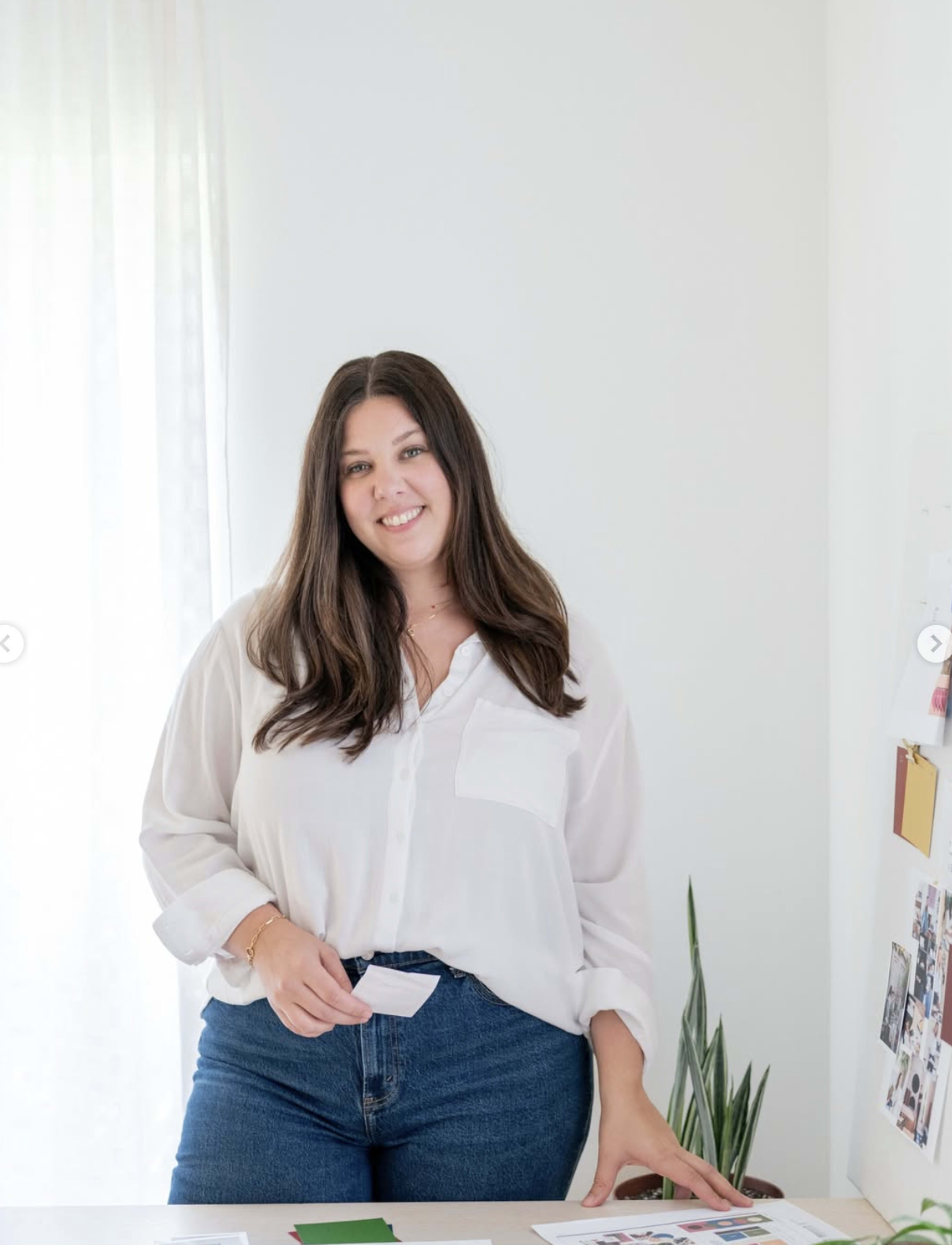 A woman in a white shirt and blue jeans stands at a desk in a bright, minimalistic room with a plant and various items on the surface.