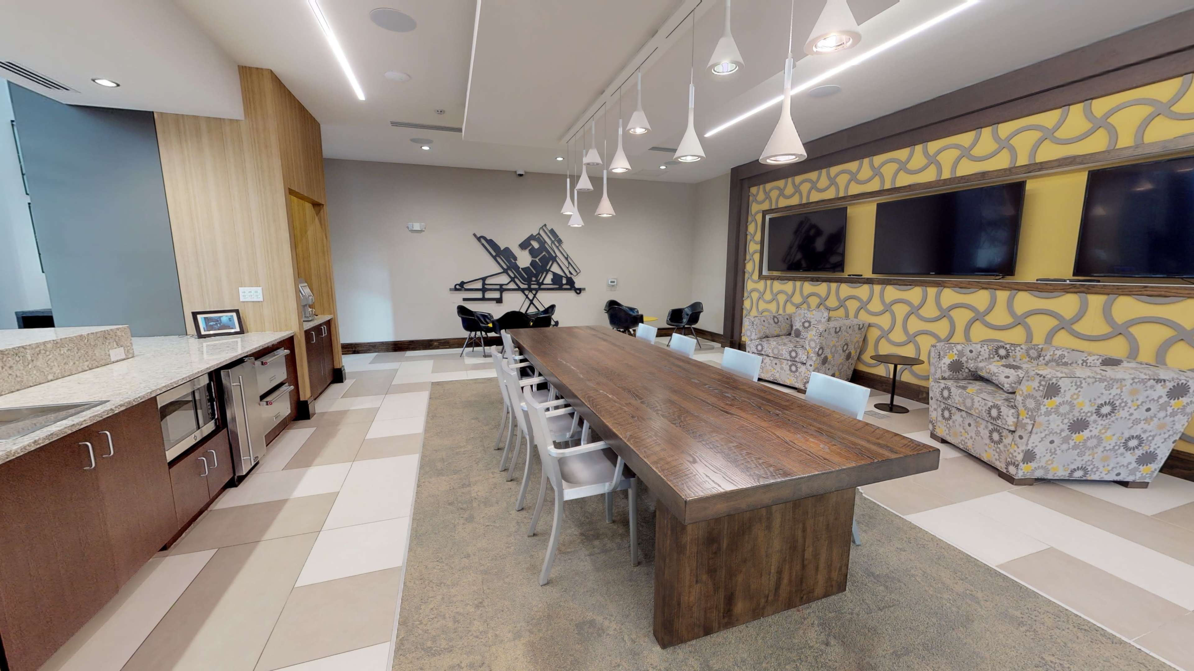 A modern communal space featuring a long wooden table, several chairs, and a patterned wall, with two large televisions mounted on one side.