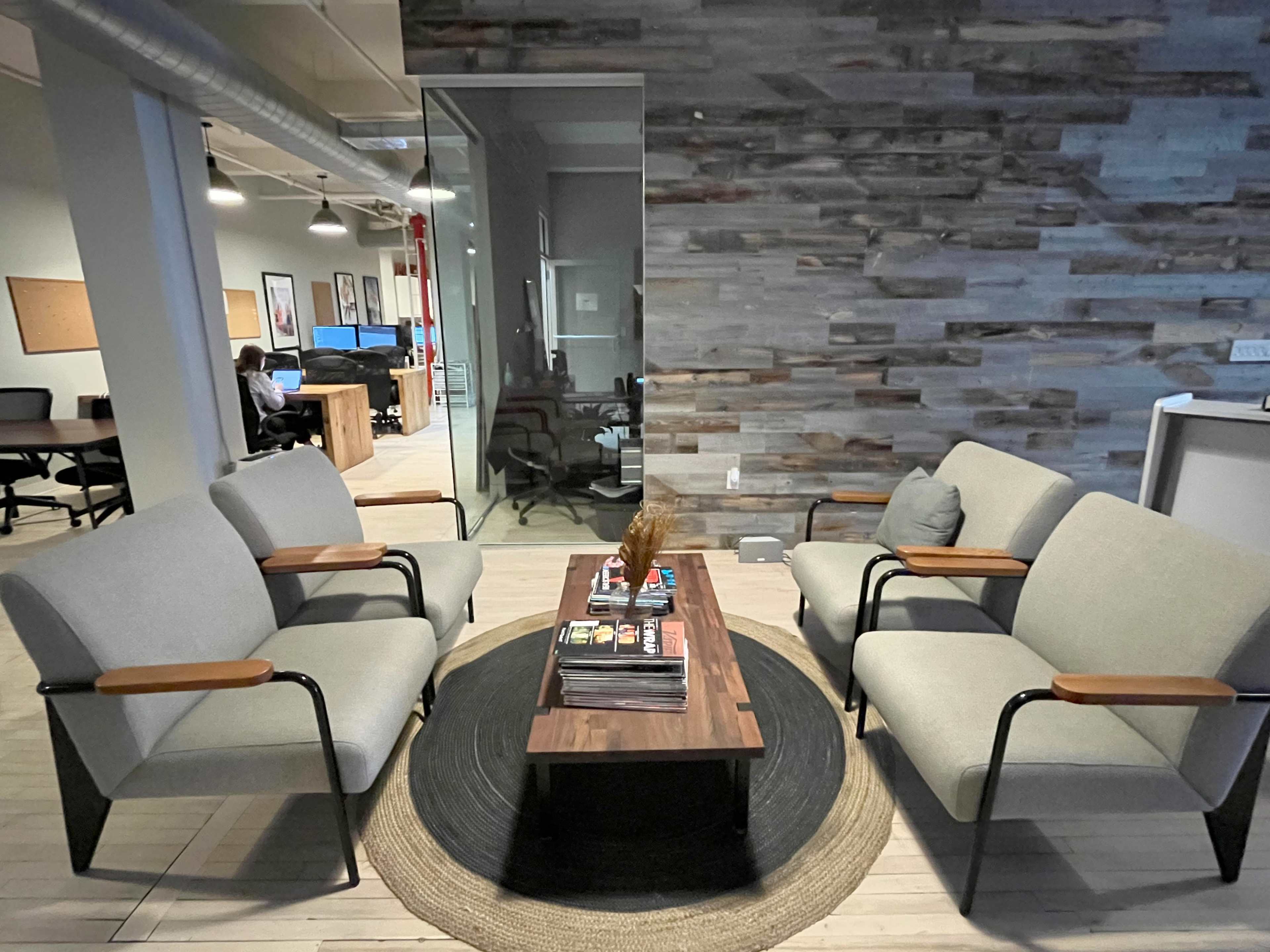 The image shows a modern office space with two upholstered couches facing a wooden coffee table, surrounded by a circular rug, and a textured stone wall in the background.