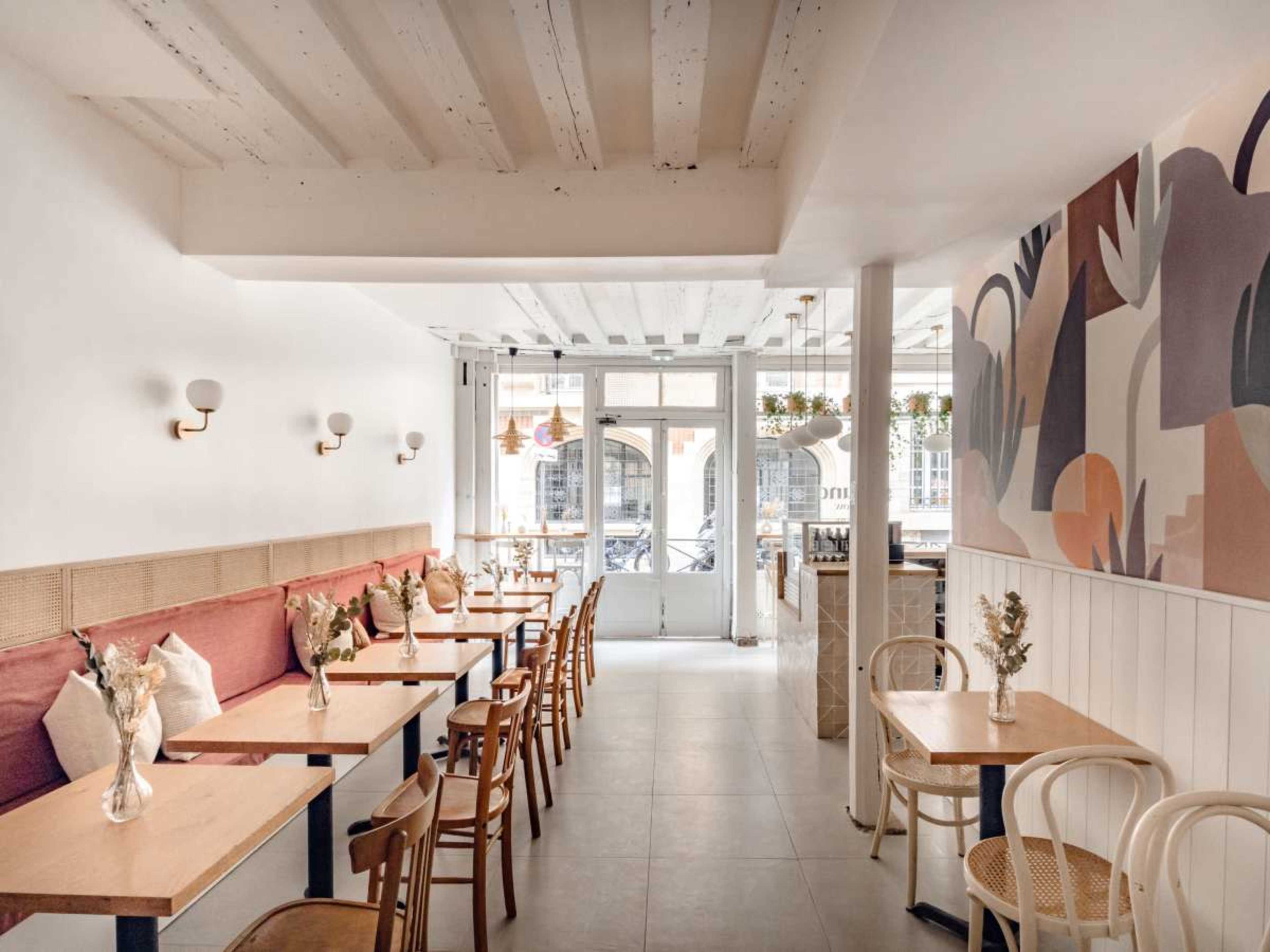 The image shows a modern café interior with light-colored wooden furniture, a mural on the wall, and large windows allowing natural light to enter.