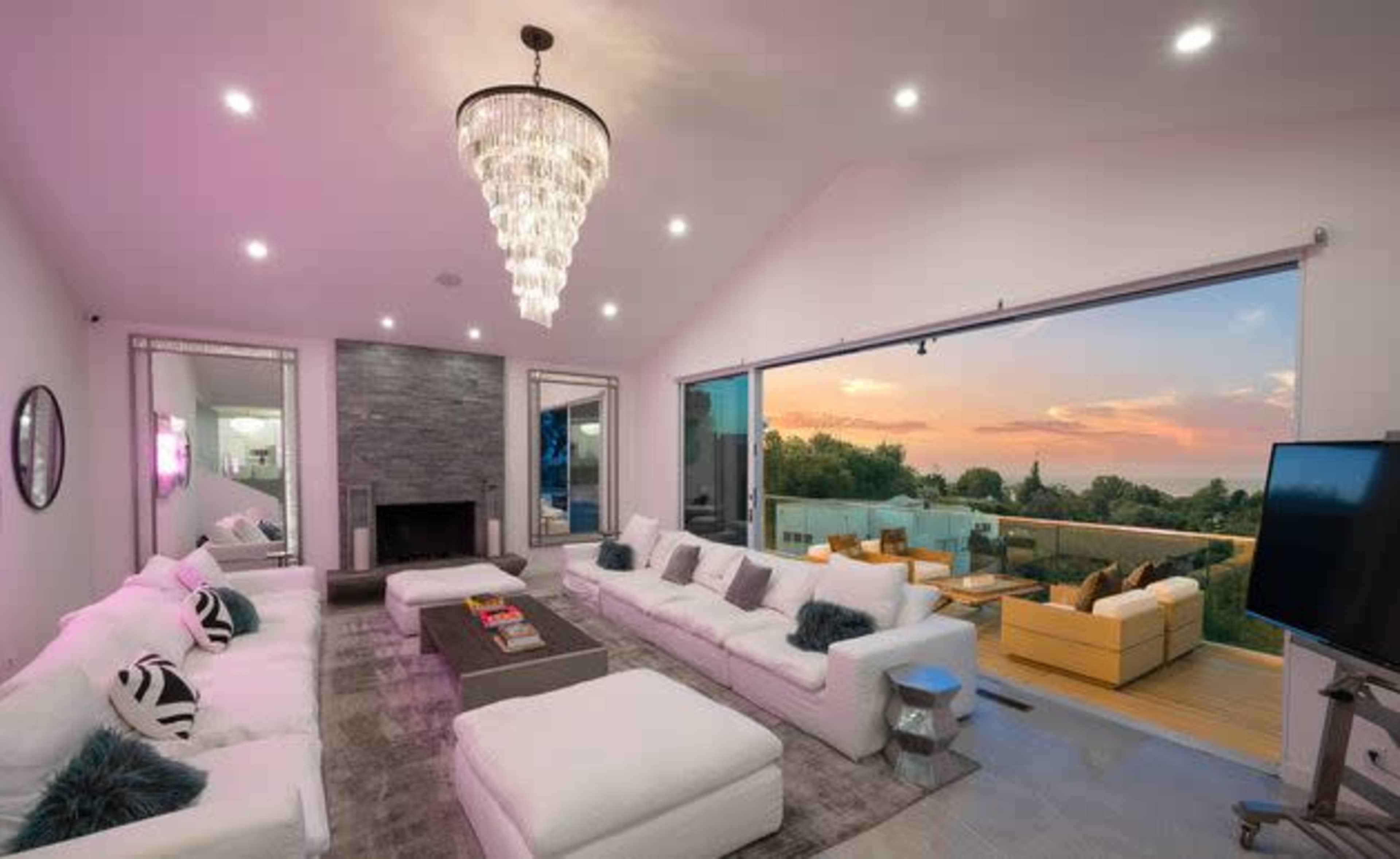 A modern living room features white sofas, a chandelier, and large glass doors opening to a balcony with a sunset view.