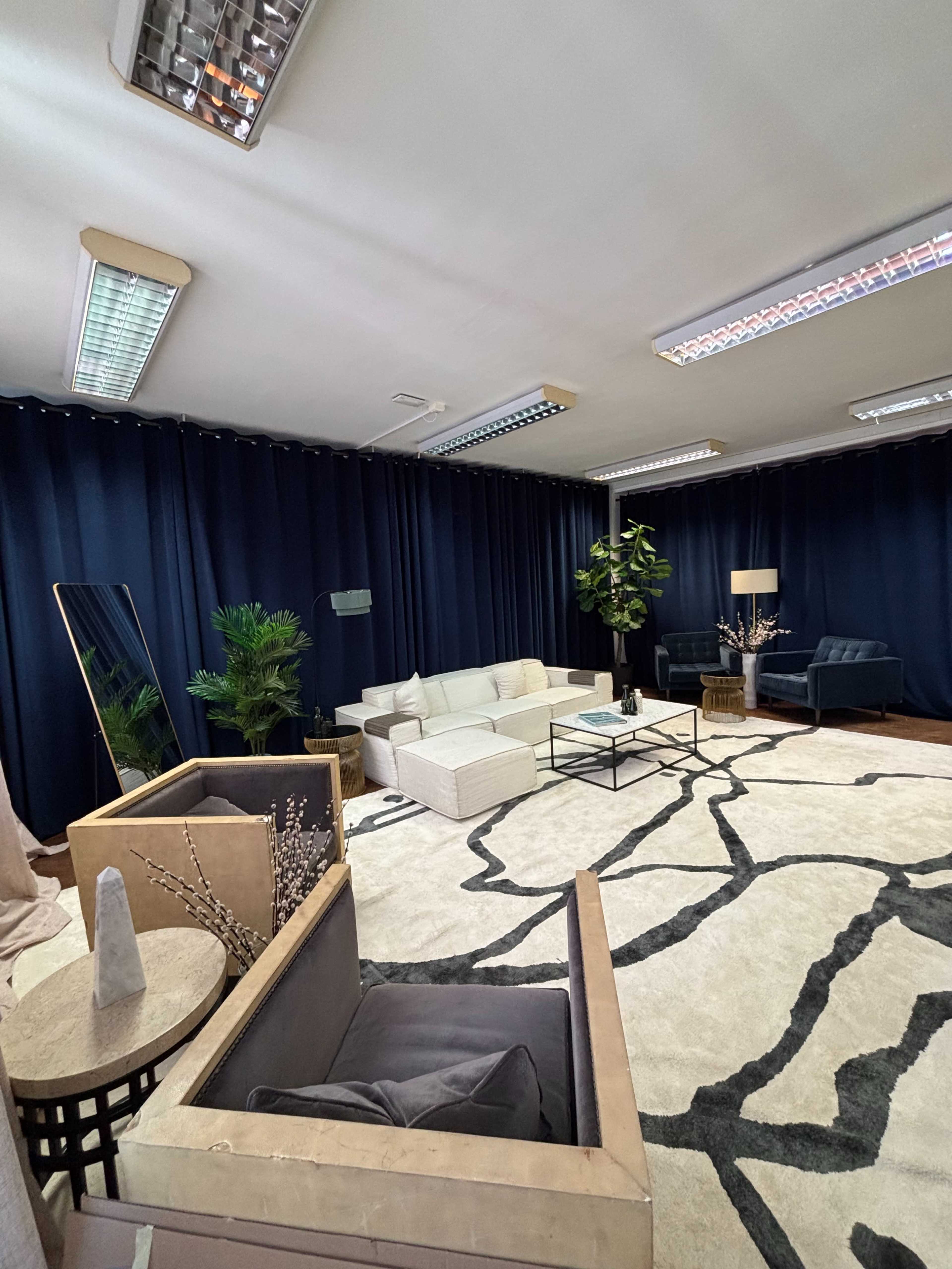The image displays a modern lounge area with a large rug, couches, a coffee table, and decorative plants, all set against dark curtains.