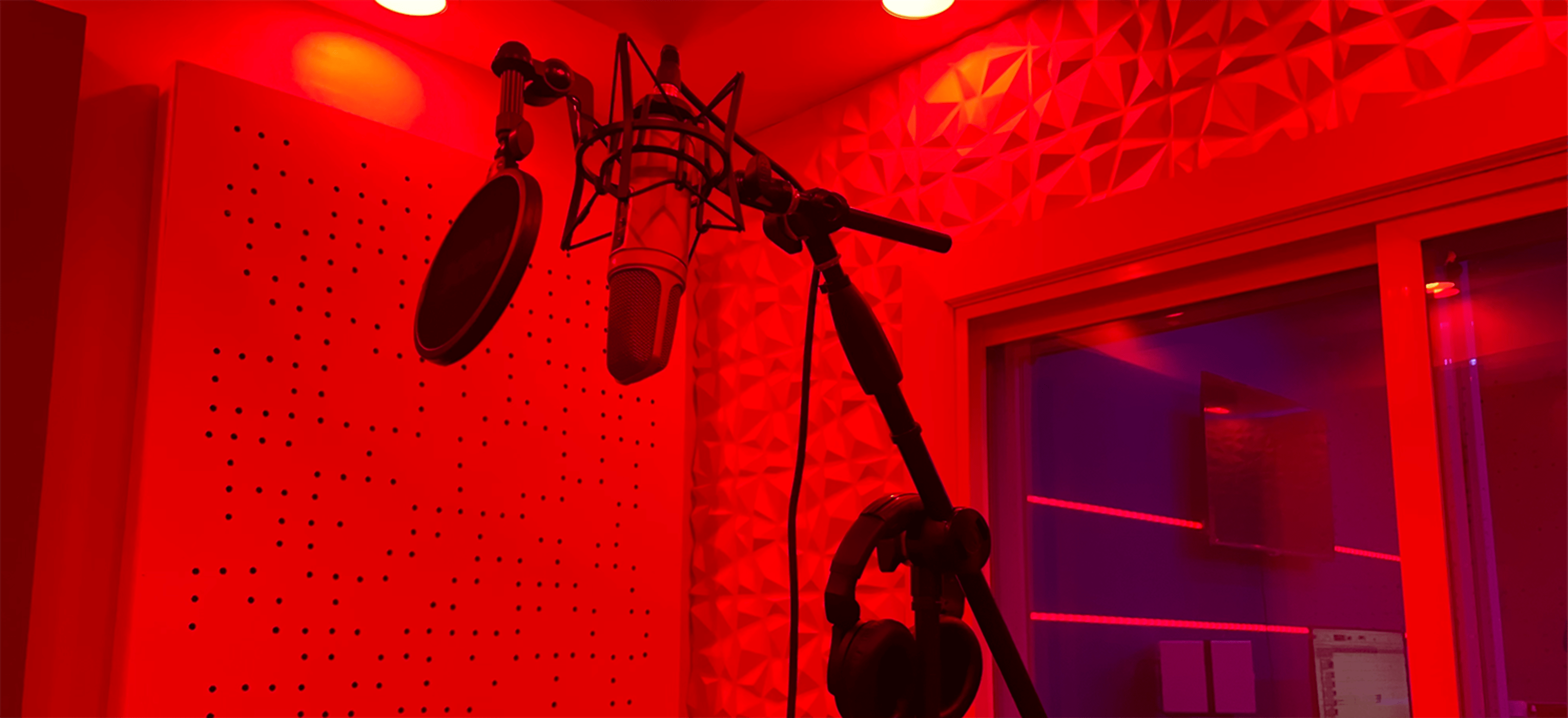 A recording studio with a microphone on a stand, headphones hanging nearby, and red lighting illuminating the walls.