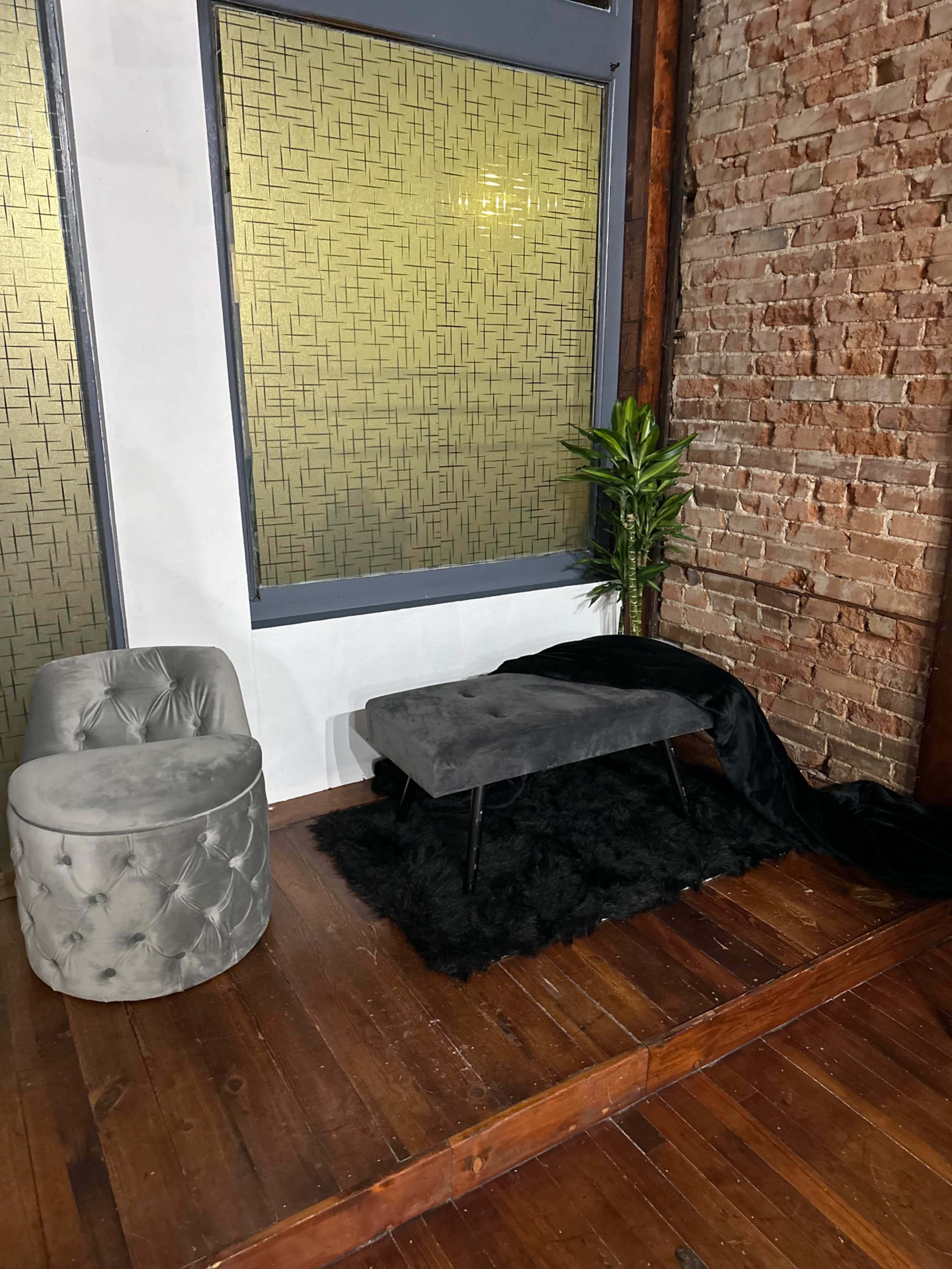 The scene features a small seating area with a plush gray chair, a fabric-covered bench, a green plant, and a dark rug on a wooden floor against a backdrop of a brick wall and decorative windows.