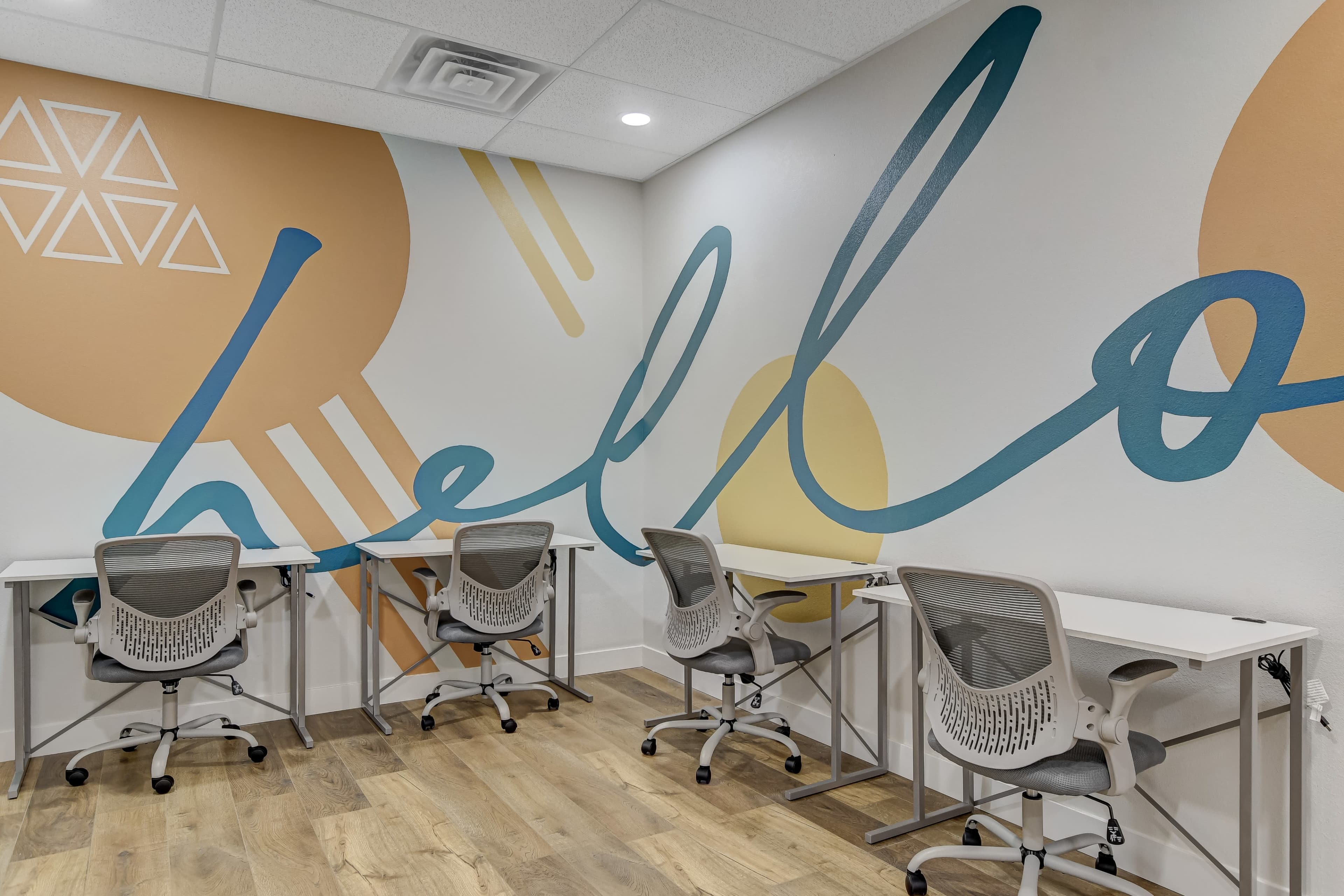 A modern workspace with four chairs and desks arranged against a wall featuring a colorful mural that includes the word "hello."