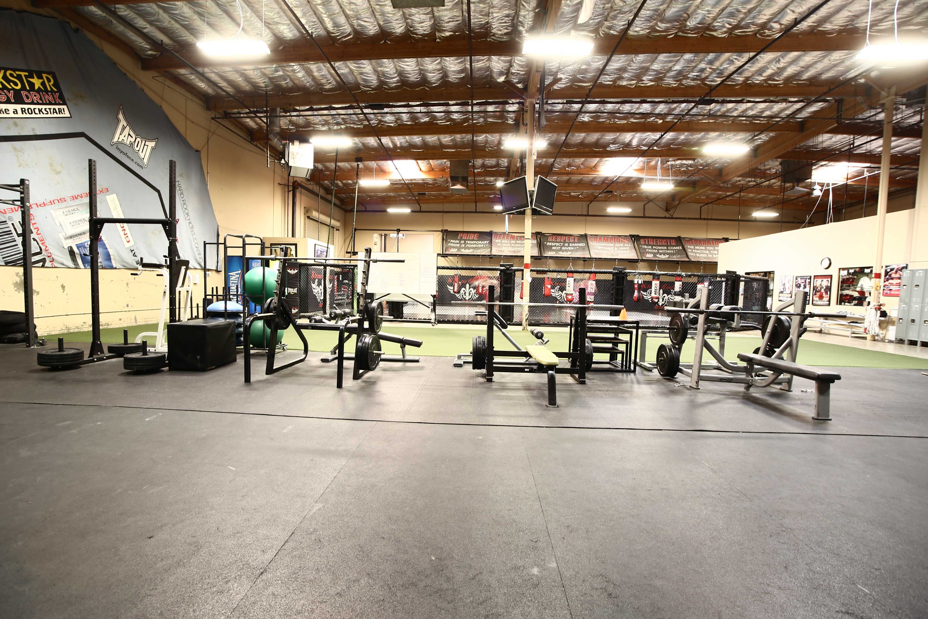A fitness gym interior featuring various weightlifting equipment arranged on a black rubber floor.