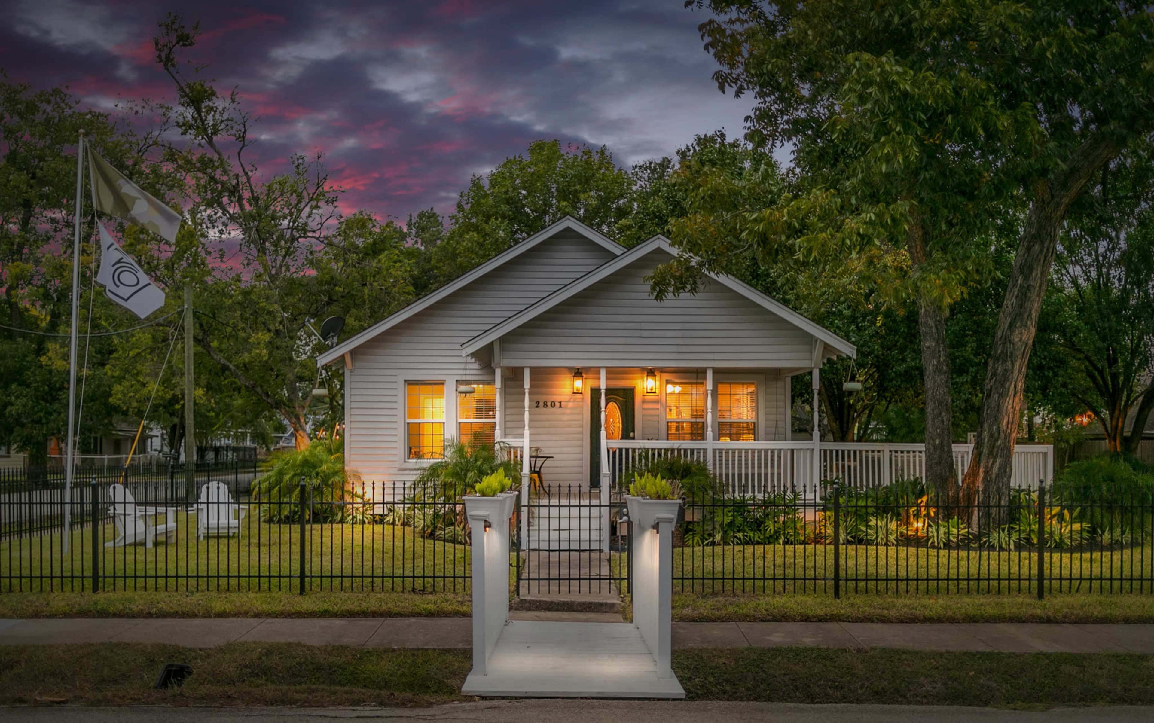 A single-story gray house with a porch is surrounded by greenery and a white picket fence under a dramatic sky at dusk.