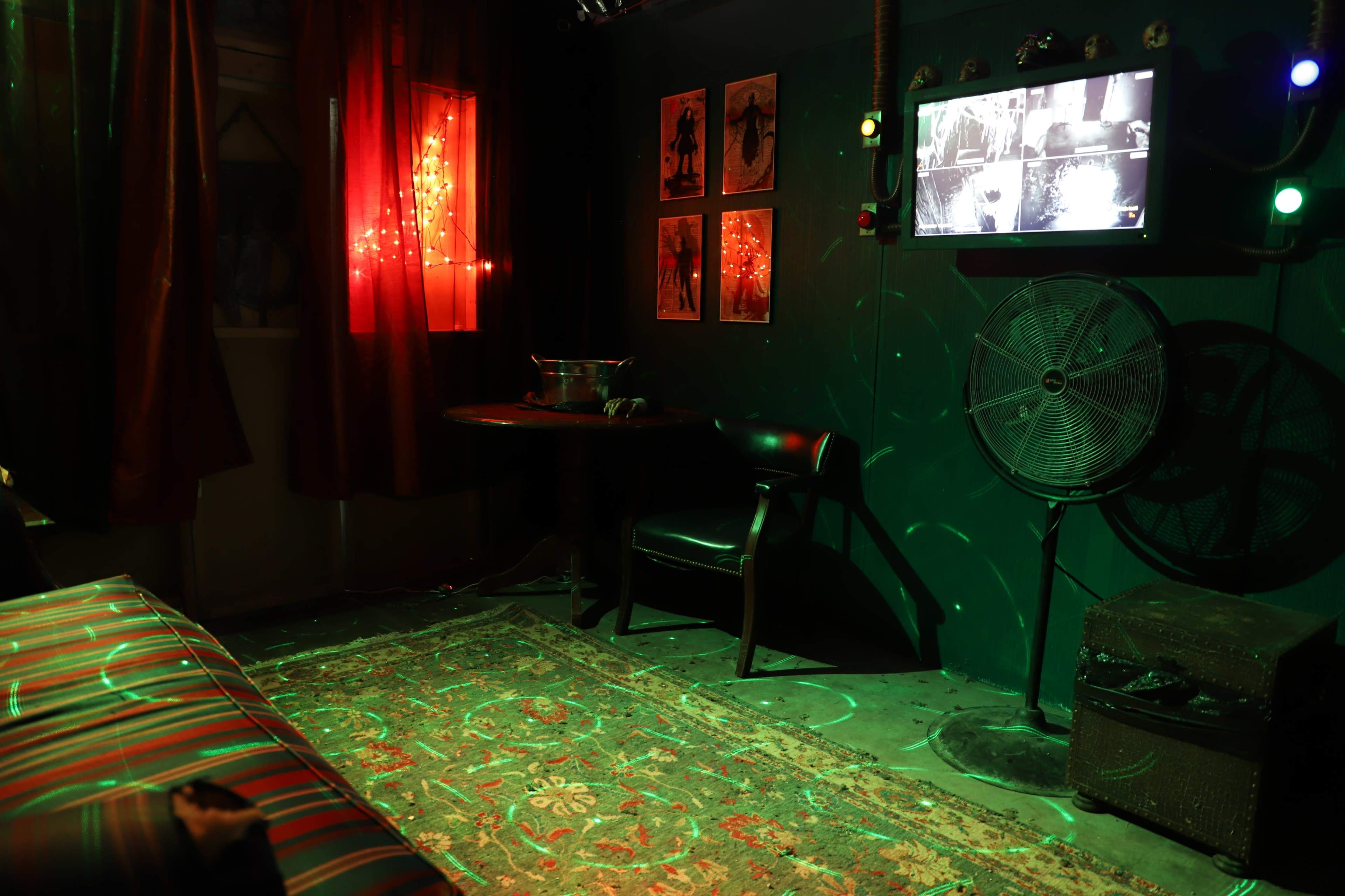 A dimly lit room features a table, a chair, a fan, and a television screen surrounded by red curtains and decorative lights.