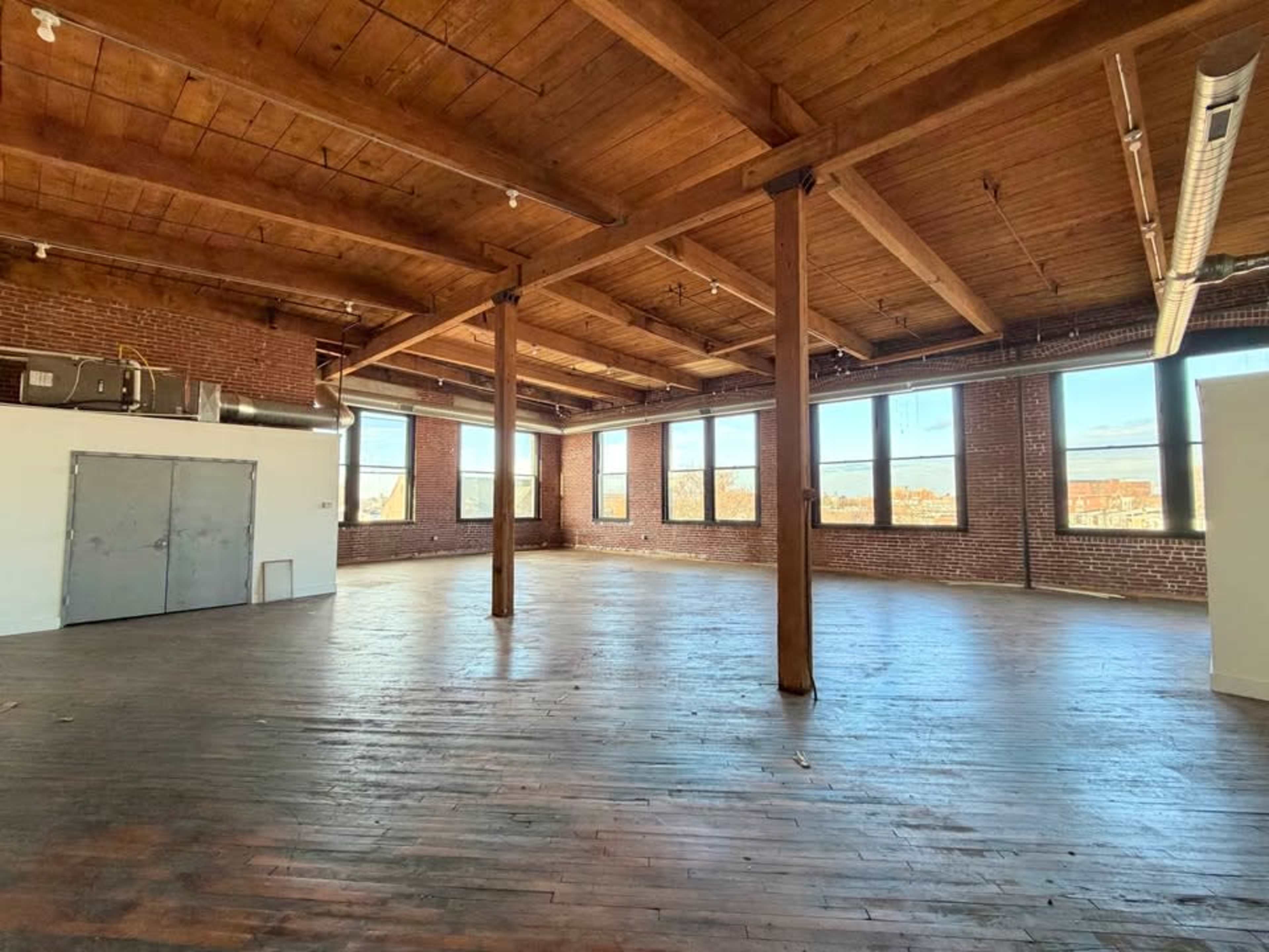 The image shows an open, spacious room with wooden floors, exposed brick walls, large windows, and wooden ceiling beams.