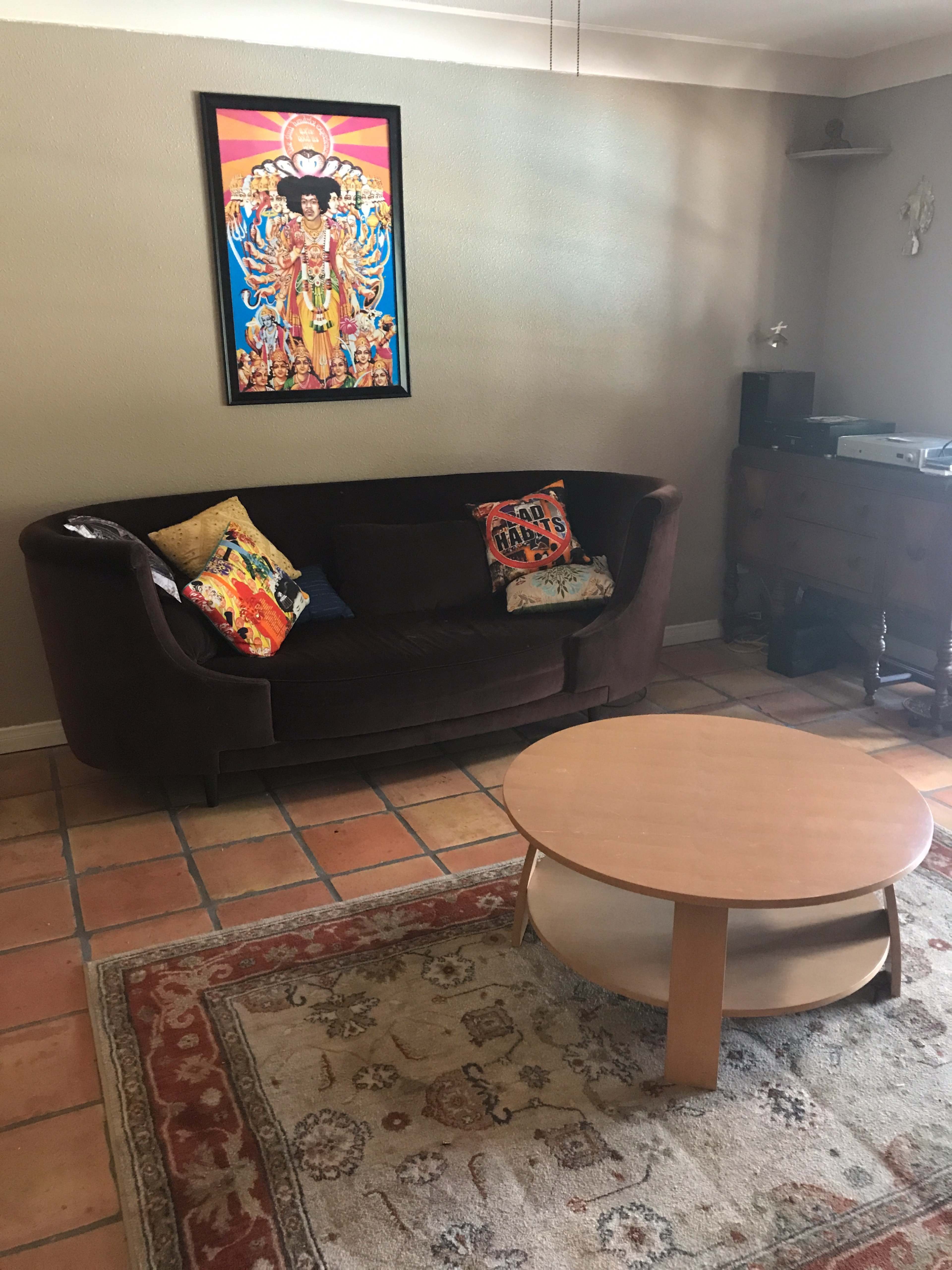 A living room with a brown couch, a round wooden coffee table, a patterned rug, and a colorful wall art piece above the sofa.