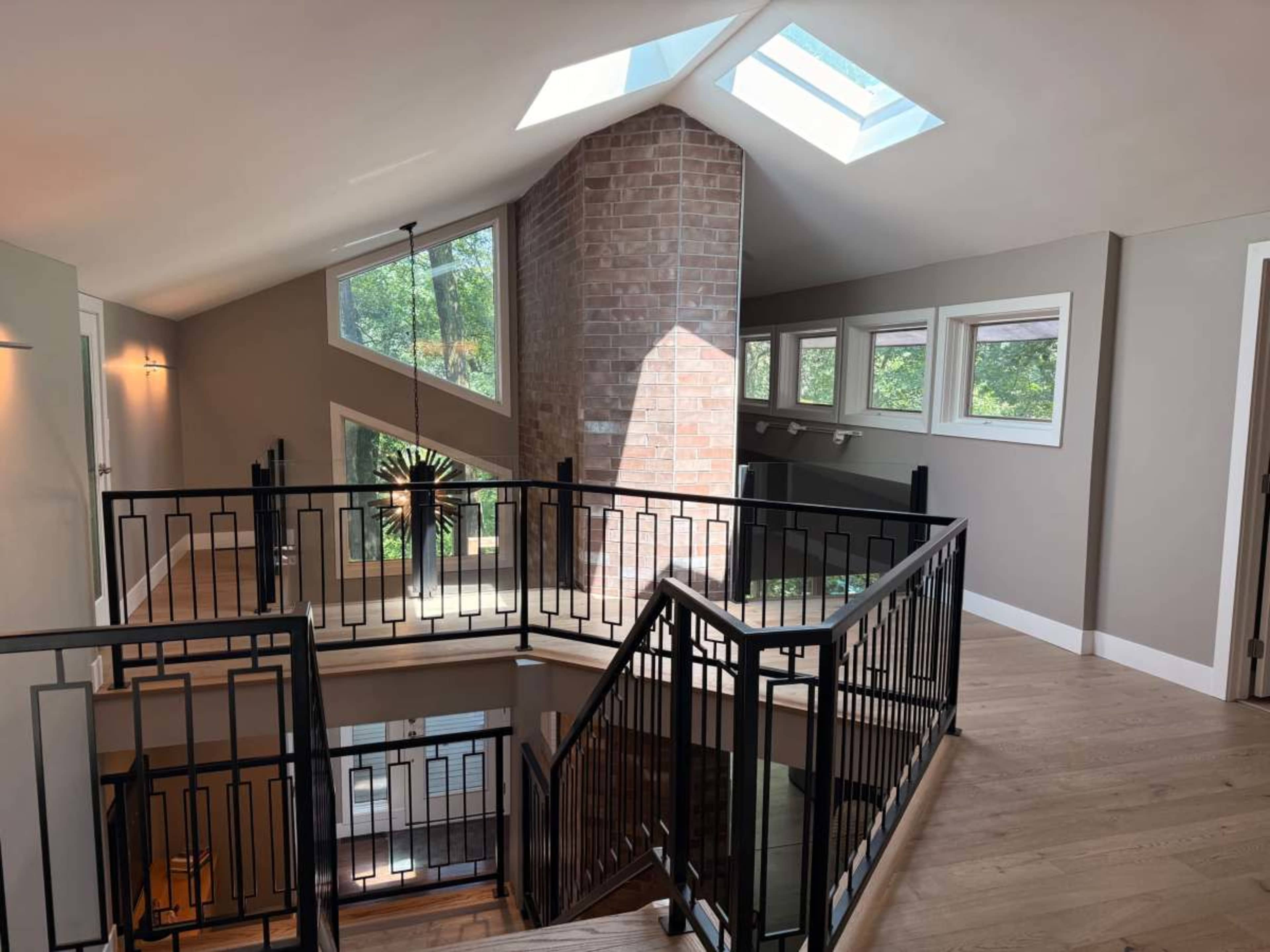 The image shows a well-lit, modern interior of a staircase area featuring large windows and a skylight, along with a brick wall accent.