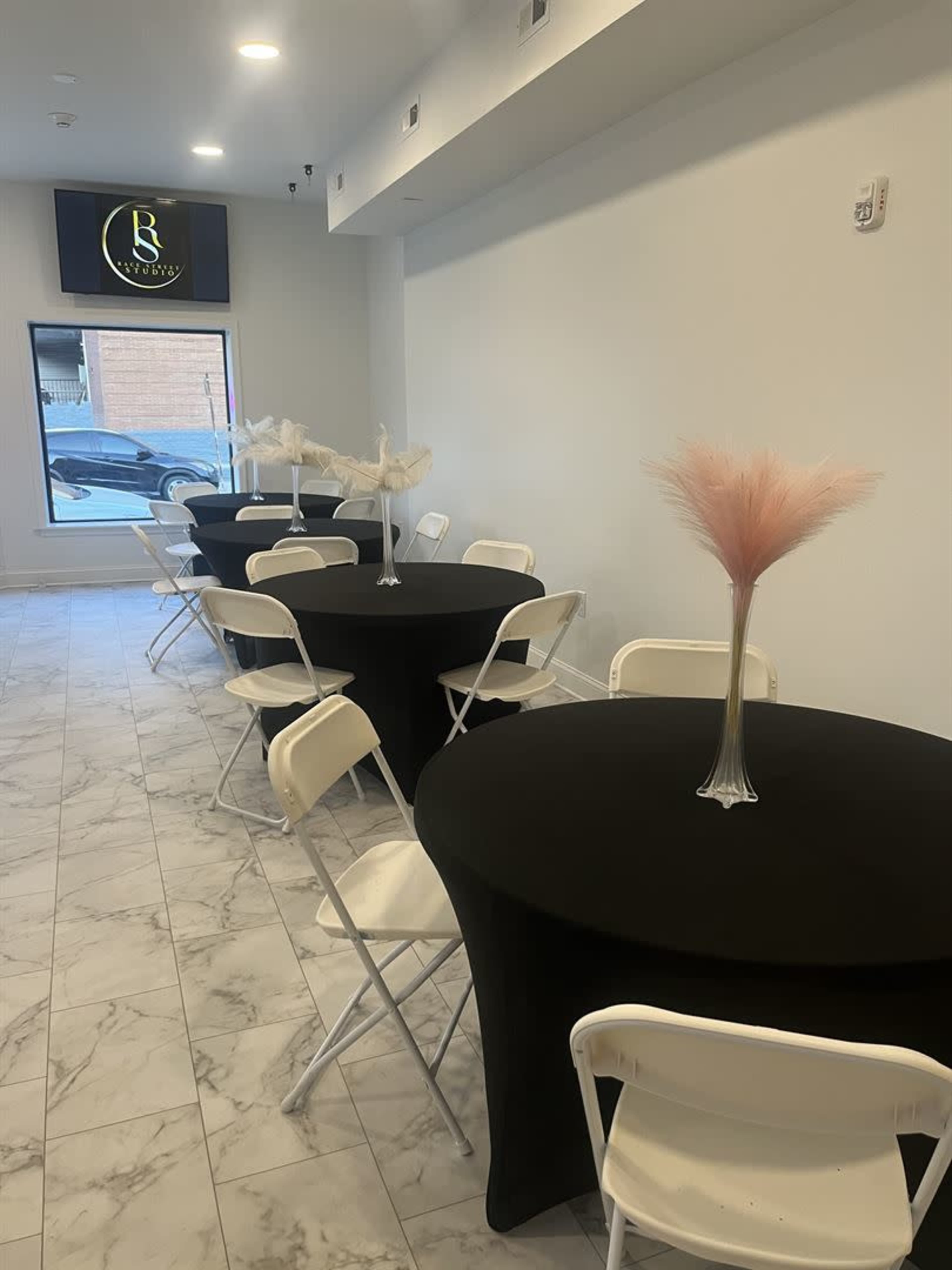 The image shows a spacious, light-filled event room featuring several round black tables with white chairs, each adorned with a decorative centerpiece.