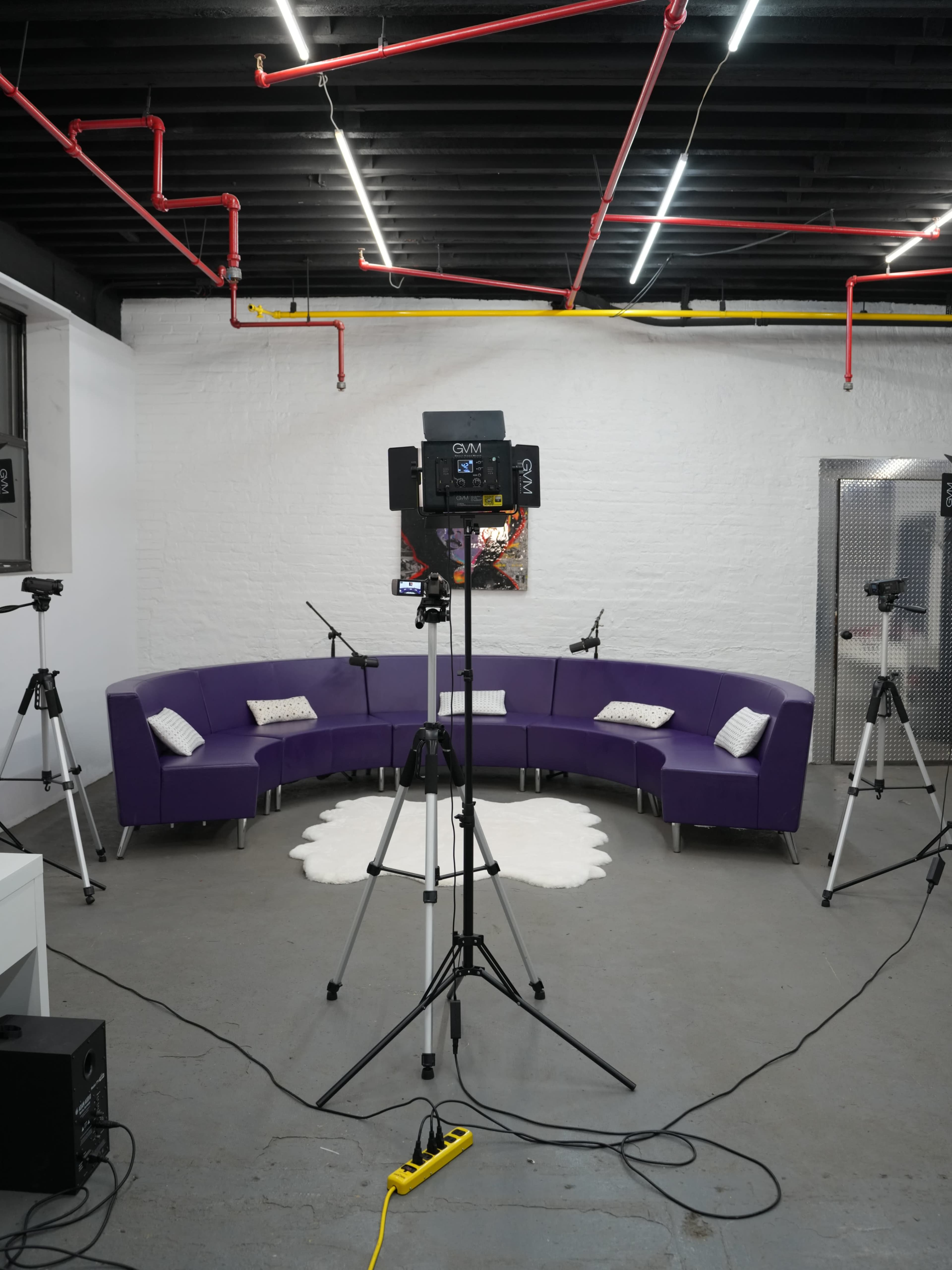 The image shows a modern studio space with a purple circular couch, a light stand, and multiple camera tripods set up around a white rug on a concrete floor.